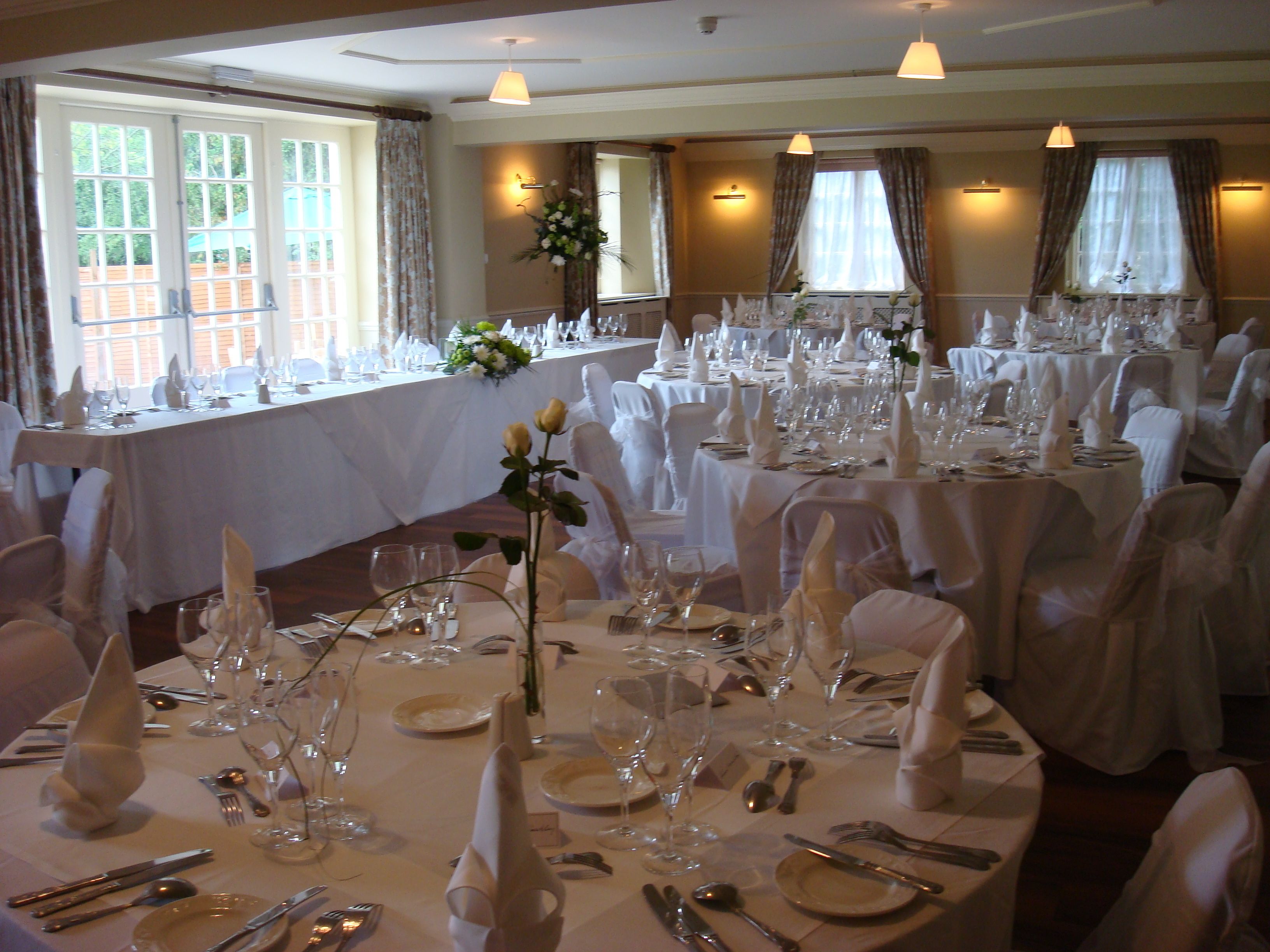 Un salón de banquetes con varias mesas redondas dispuestas para un evento formal, con manteles blancos, cristalería y sillas.
