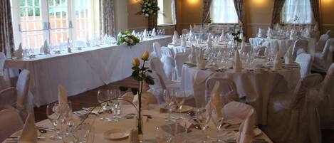 Un salón de banquetes con varias mesas redondas dispuestas para un evento formal, con manteles blancos, cristalería y sillas.
