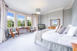 A bedroom with a large bed, a desk, two chairs, a television, and a view of greenery through the windows. - Leigh Park Country House Hotel & Vineyard, BW Signature Collection (Bradford-on-Avon)