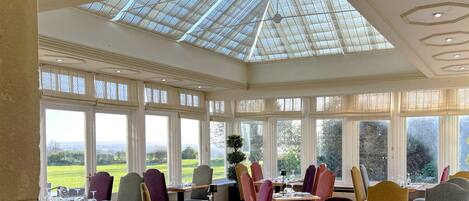 A dining area with large windows, a glass ceiling, and a view of a green landscape.