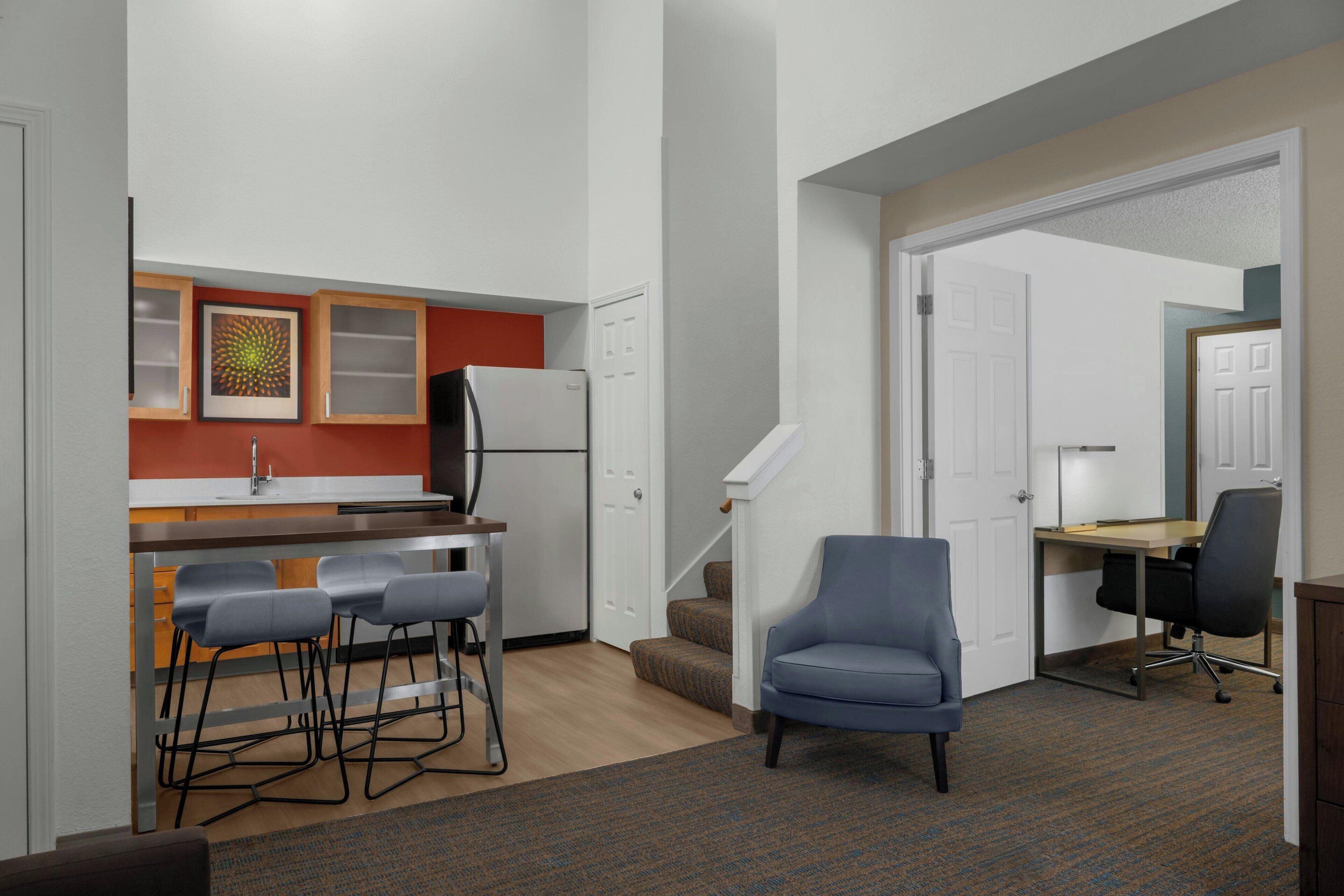 Um apartamento compacto com cozinha americana, área de refeições com banquetas, uma poltrona azul e uma escrivaninha com cadeira.