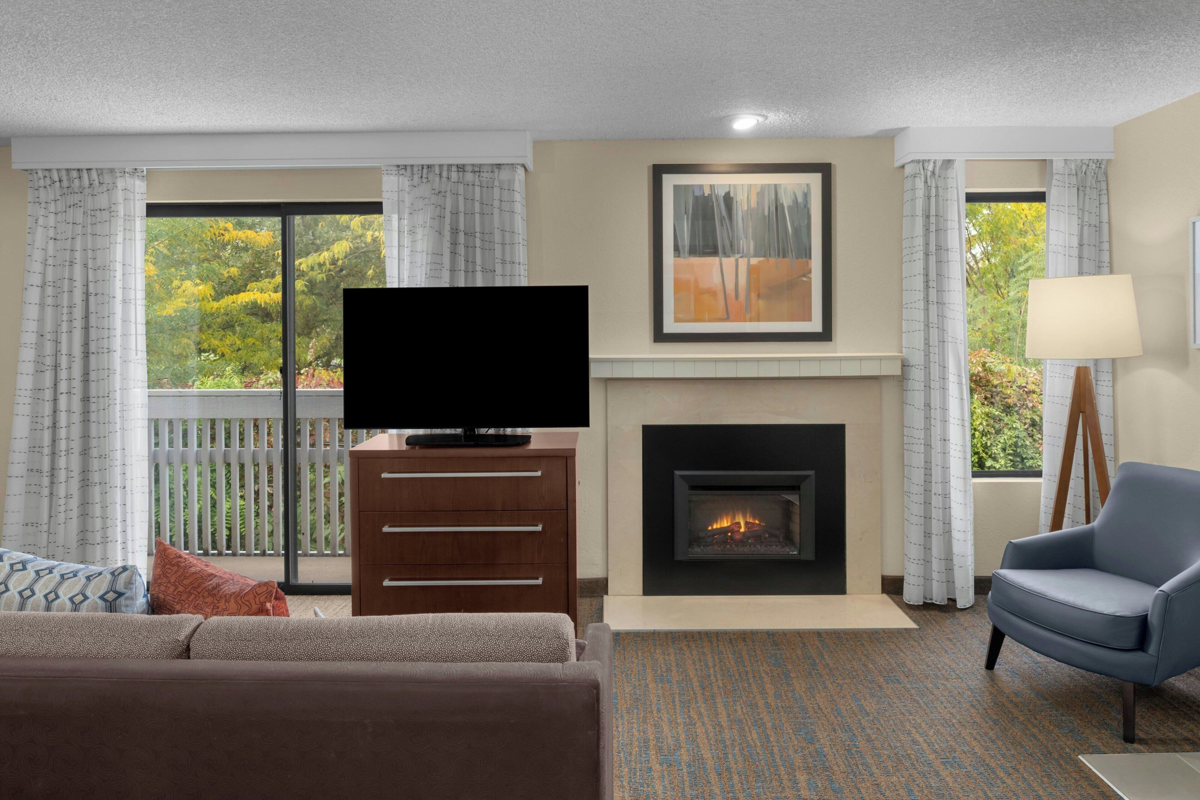 A living room with a fireplace, a television, a sofa, a chair, and a view of trees through large windows.