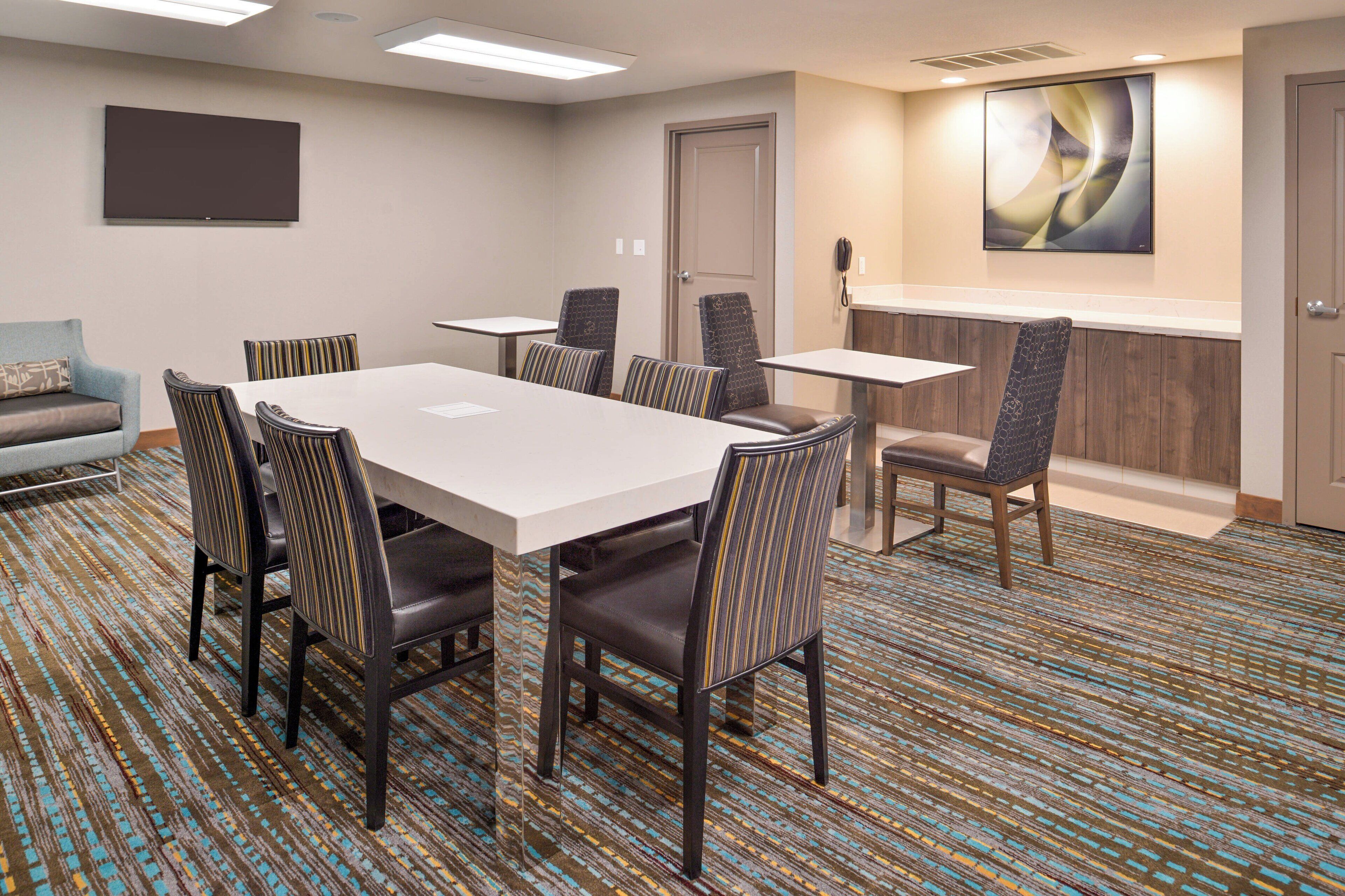 A conference room with a large table, six chairs, a TV, a phone, and a door.