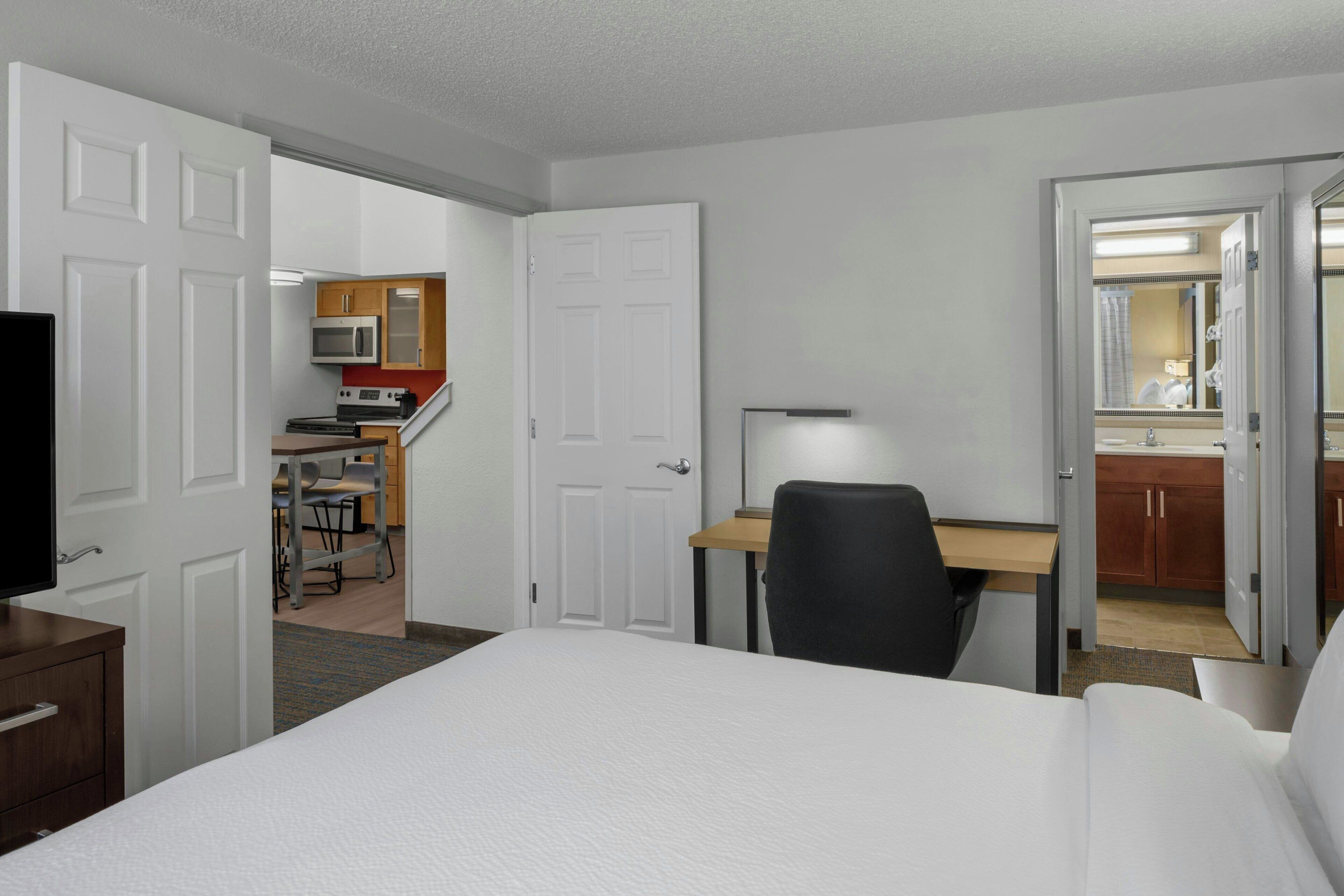 A hotel room with a bed, desk, chair, kitchenette, and a bathroom with a mirror.