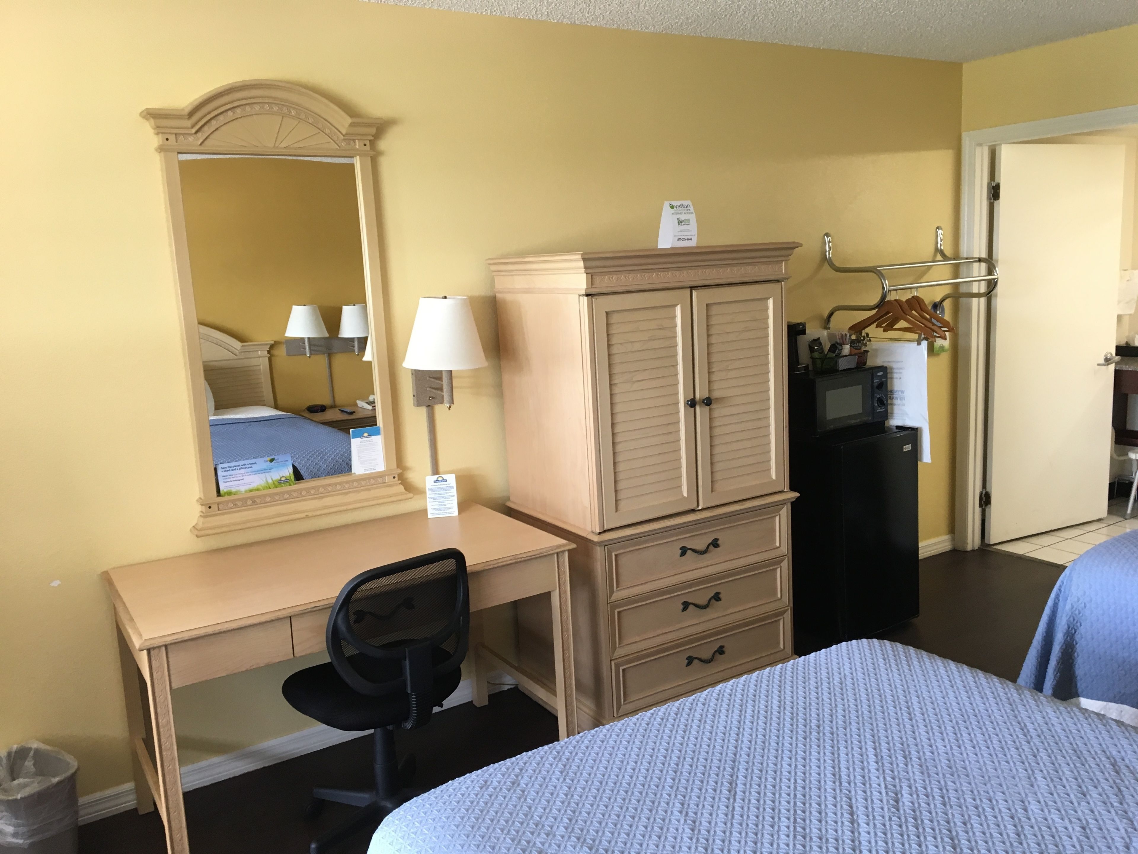 Une chambre d’hôtel avec un bureau en bois, un miroir, une armoire, un lit, une chaise et une petite kitchenette.