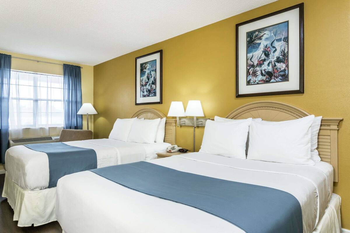 A hotel room with two beds, a desk, and a chair. There are two wall-mounted lamps and a painting above the beds.