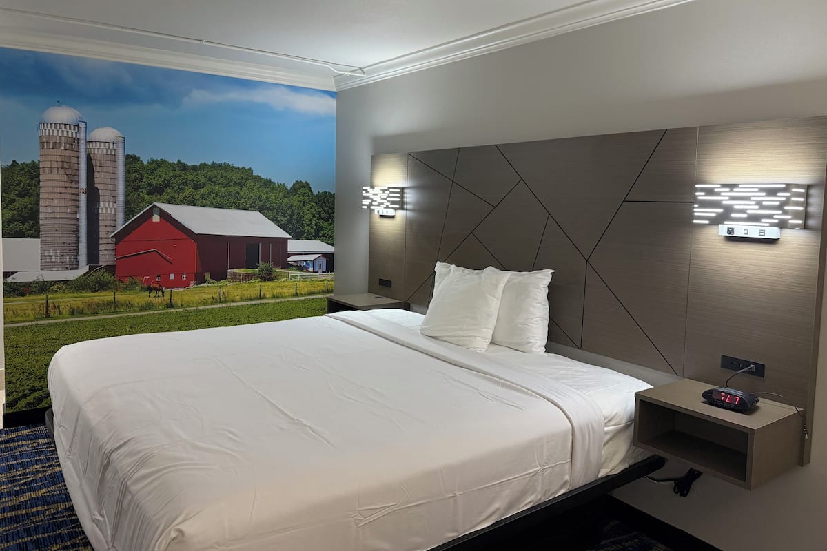 A hotel room with a large bed, a nightstand, a wall-mounted light, and a mural of a farm scene.