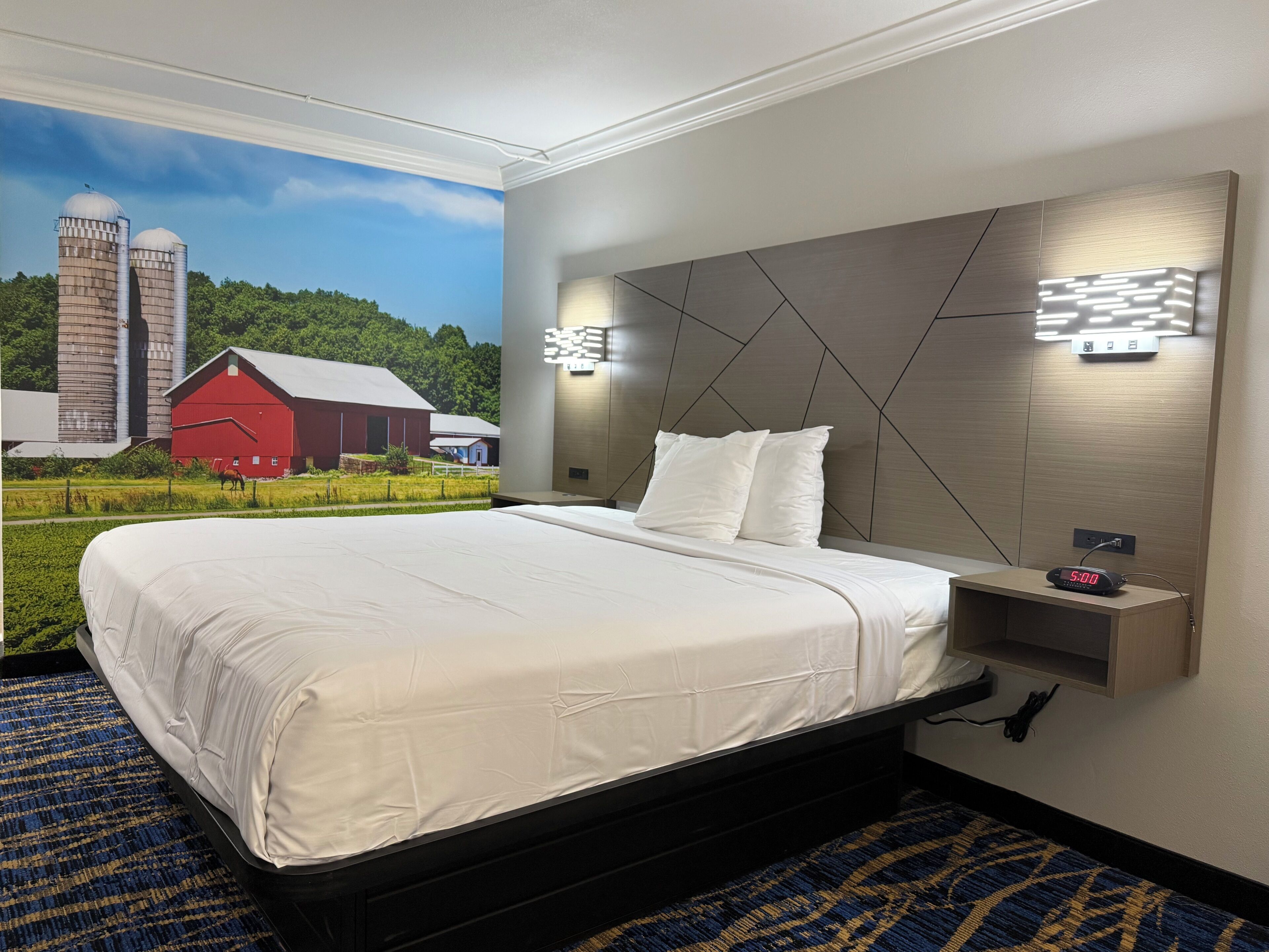 A hotel room with a large bed, a headboard with geometric patterns, a bedside table with a clock, and a mural of a farm scene.