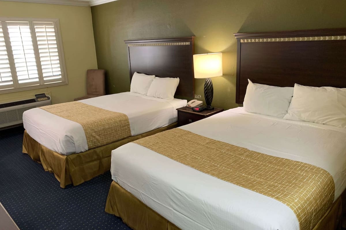 A hotel room with two beds, a nightstand with a lamp, and a window with blinds.