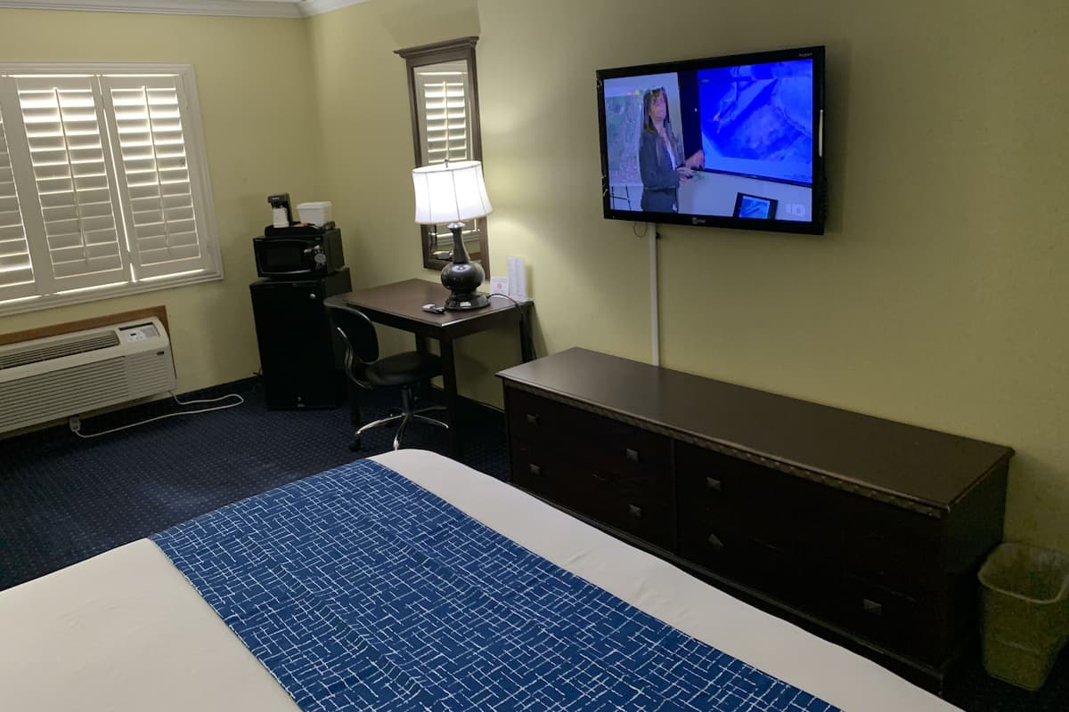 A hotel room with a large bed, a desk with a lamp, a television mounted on the wall, and a waste bin.