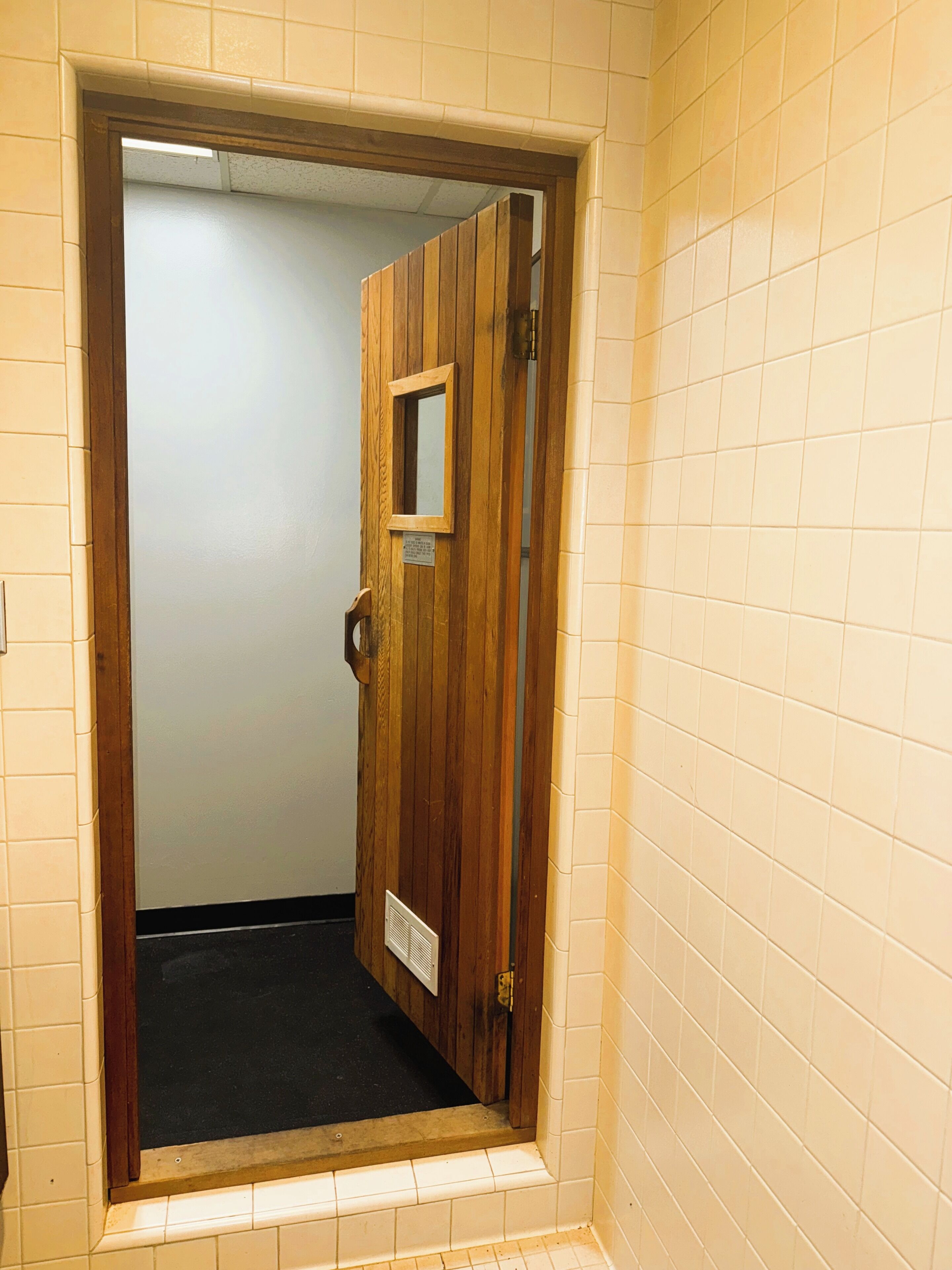 Puerta de madera con un espejo pequeño y una rejilla de ventilación, que da a un pasillo con baldosas.