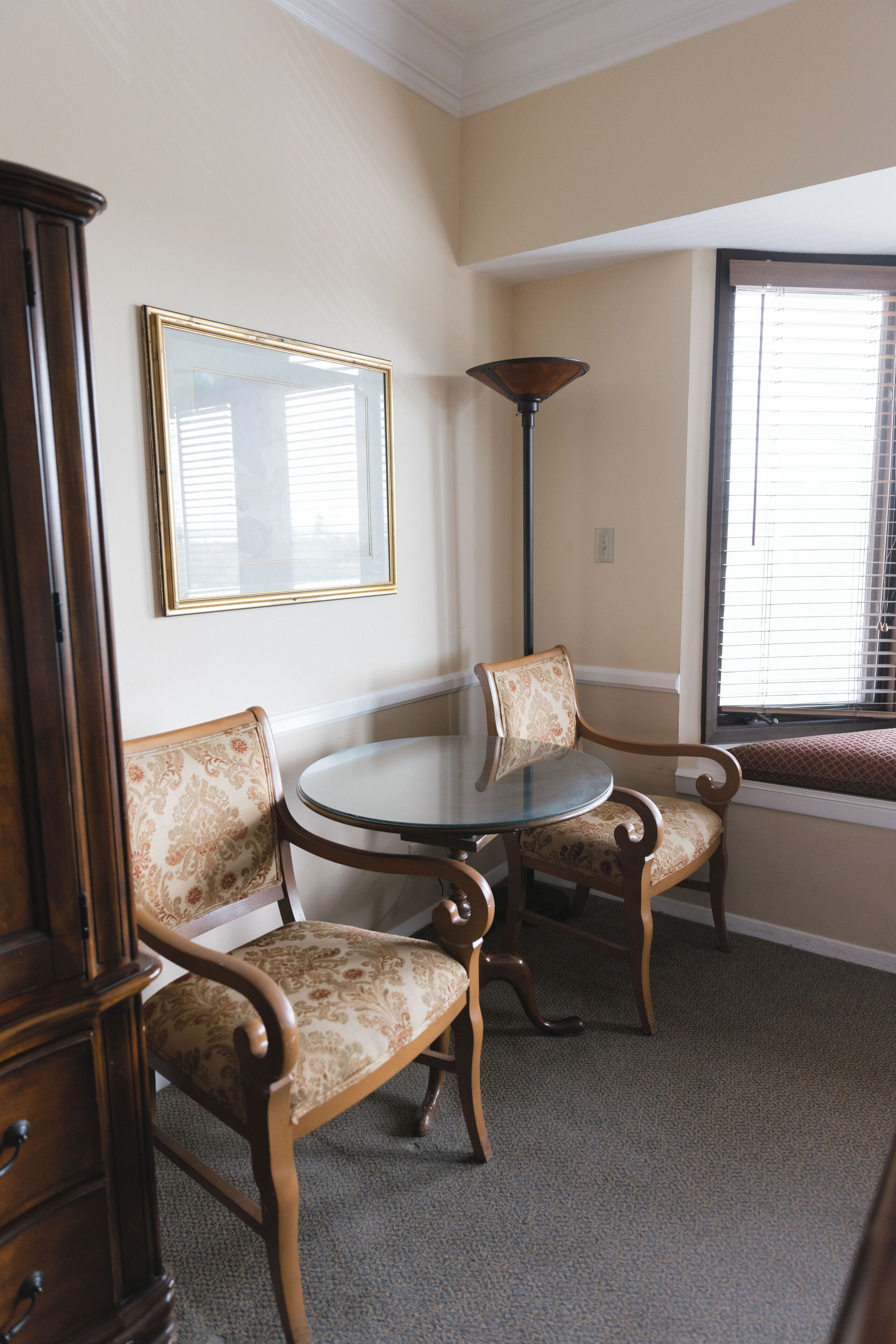 A corner room with a round glass table, two patterned chairs, a cabinet, a framed picture, and a window with blinds.