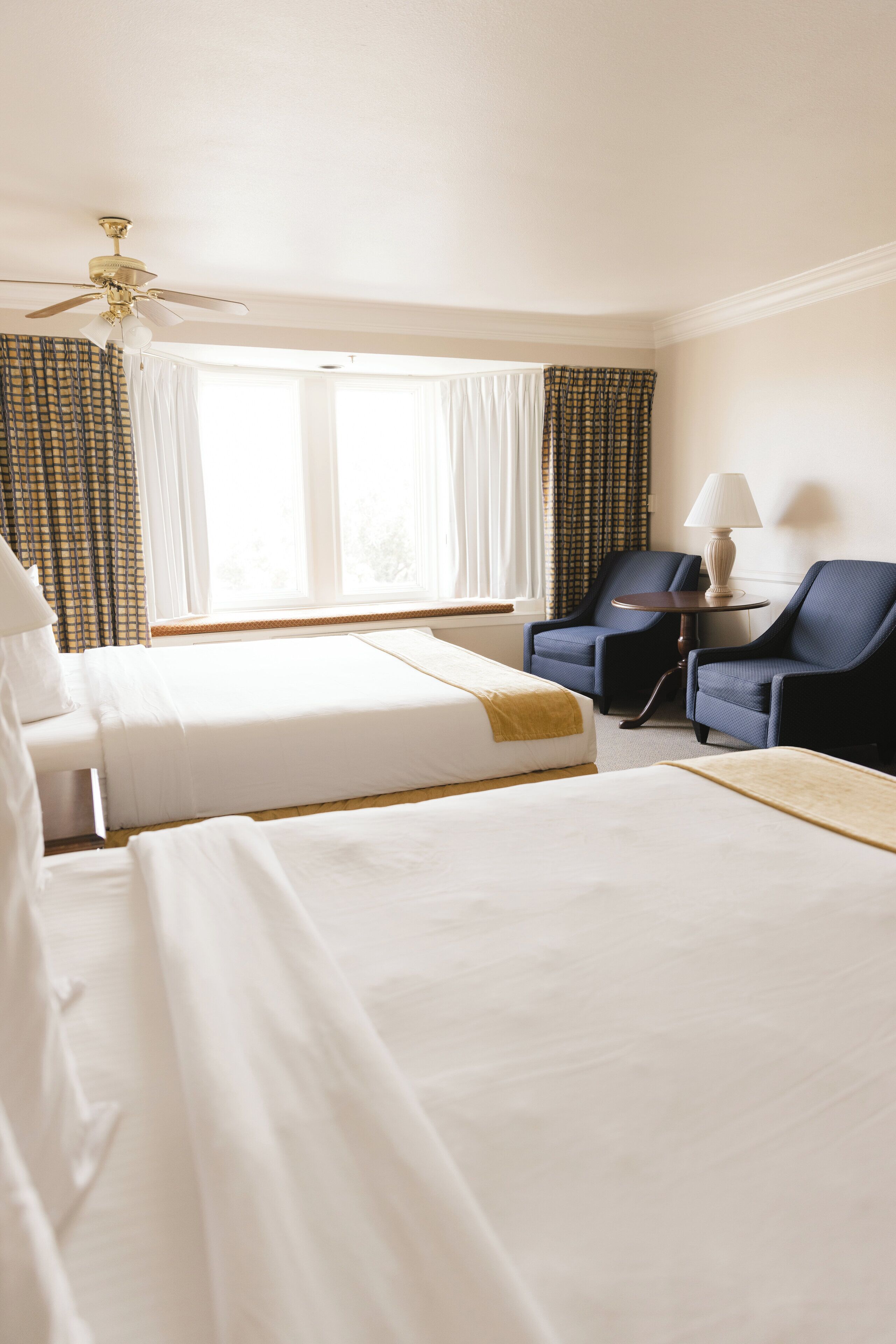 A hotel room with two beds, a desk, two chairs, a lamp, and a ceiling fan.
