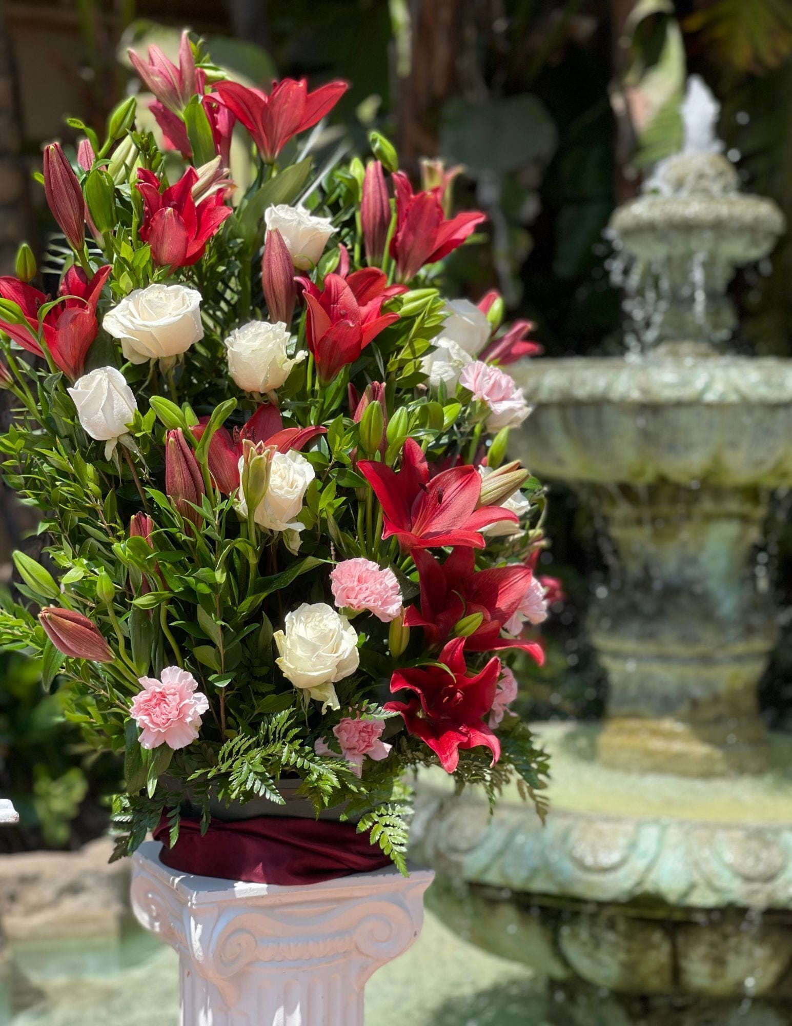 Un arrangement floral en pot, composé de fleurs rouges et blanches, est placé sur un piédestal blanc, avec une fontaine en arrière-plan.