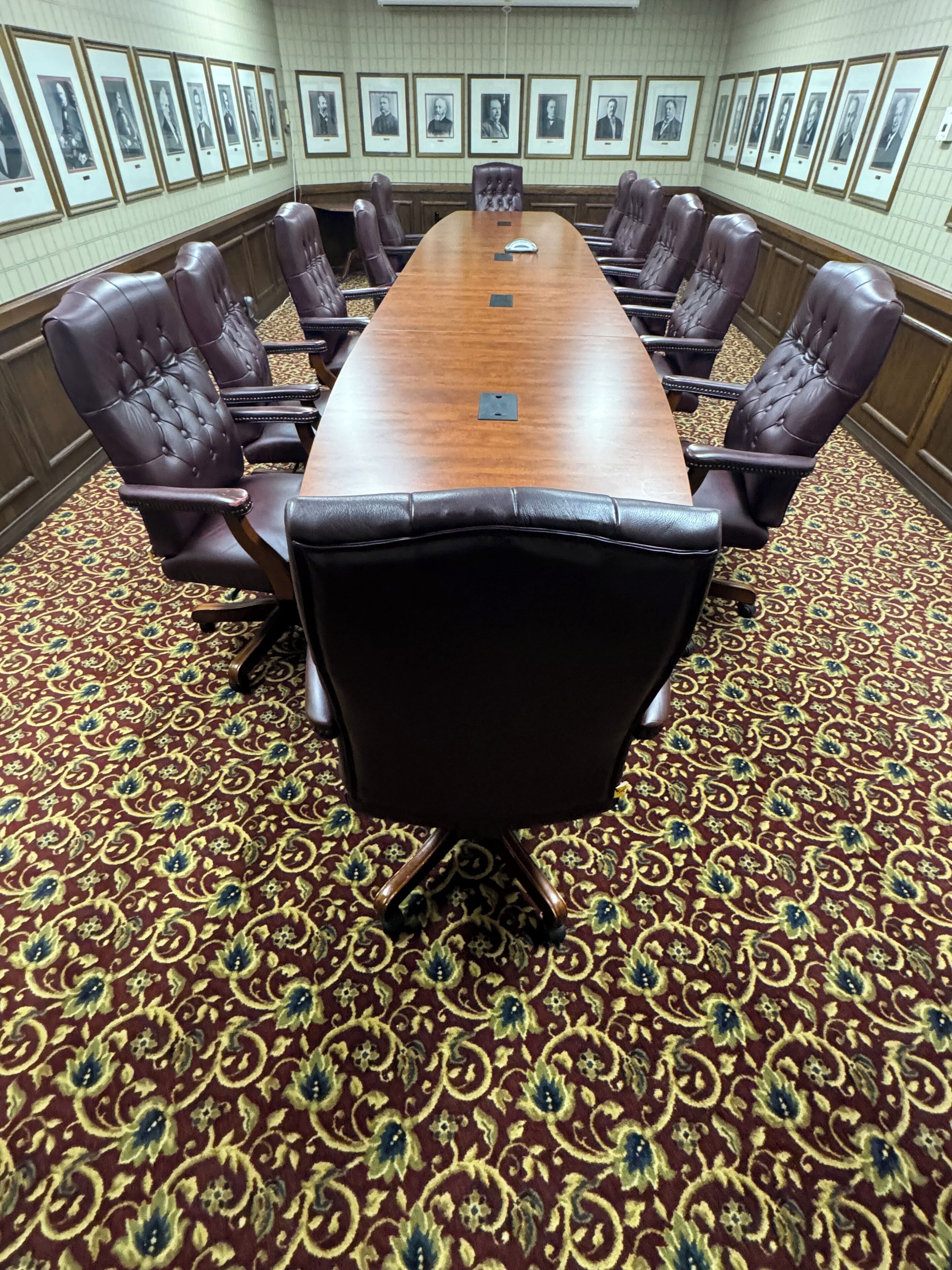 A conference room with a long table surrounded by leather chairs, framed portraits on the wall, and a patterned carpet.