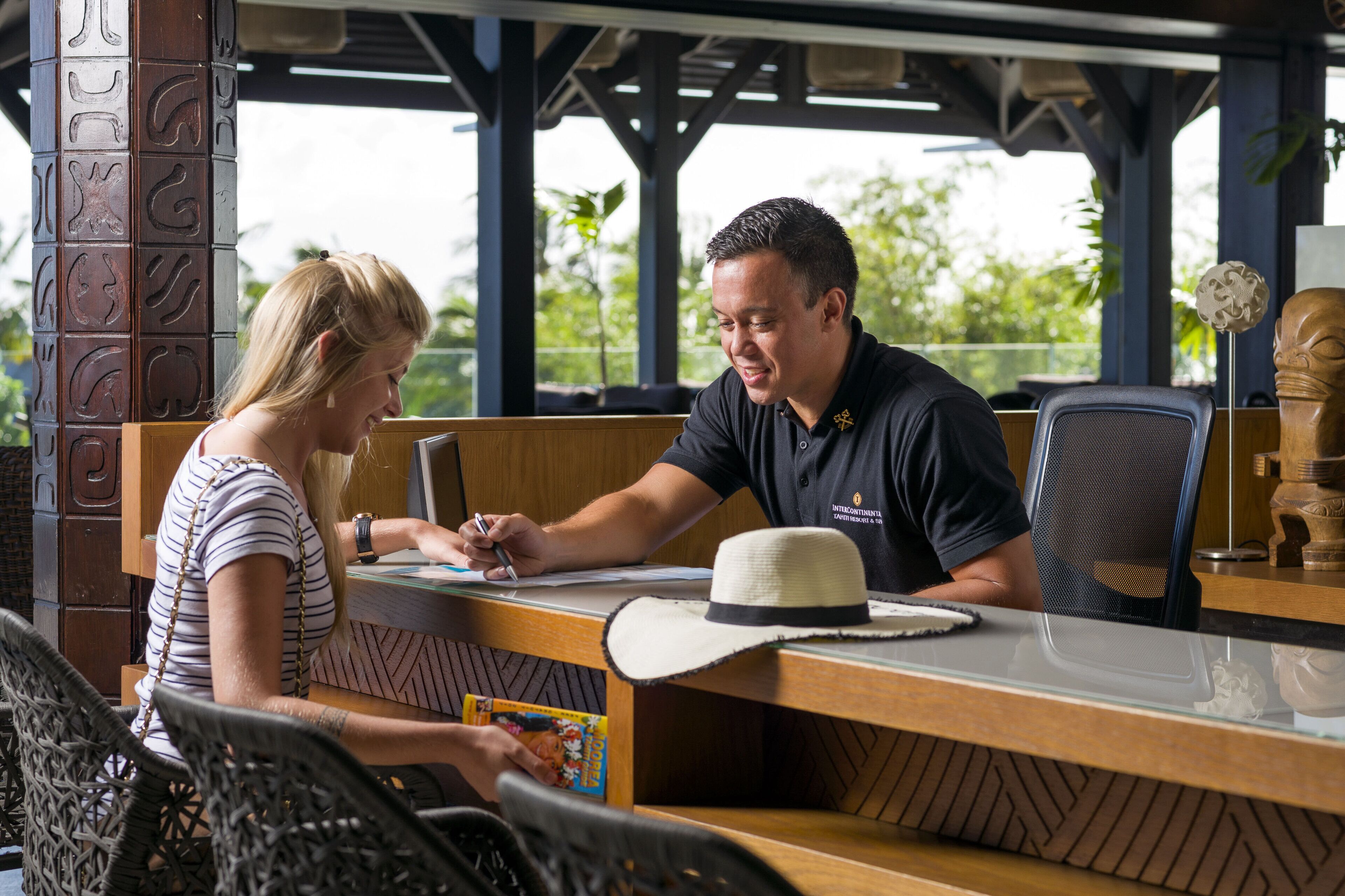A reception desk with a wooden counter, a hat on the desk, and a person seated at the desk.