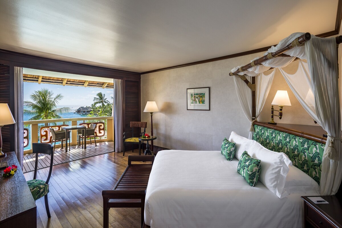 Une chambre d’hôtel avec un lit, une table et des chaises, un balcon donnant sur la mer et un tableau accroché au mur.