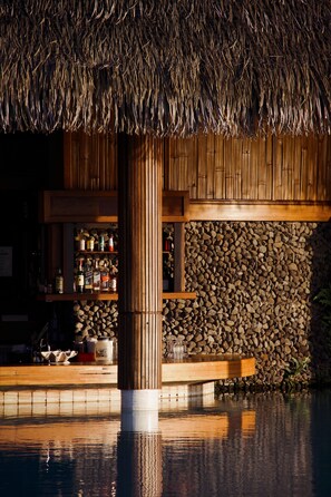 A thatched-roof structure with a wooden bar and shelves of bottles, surrounded by a stone wall and water.