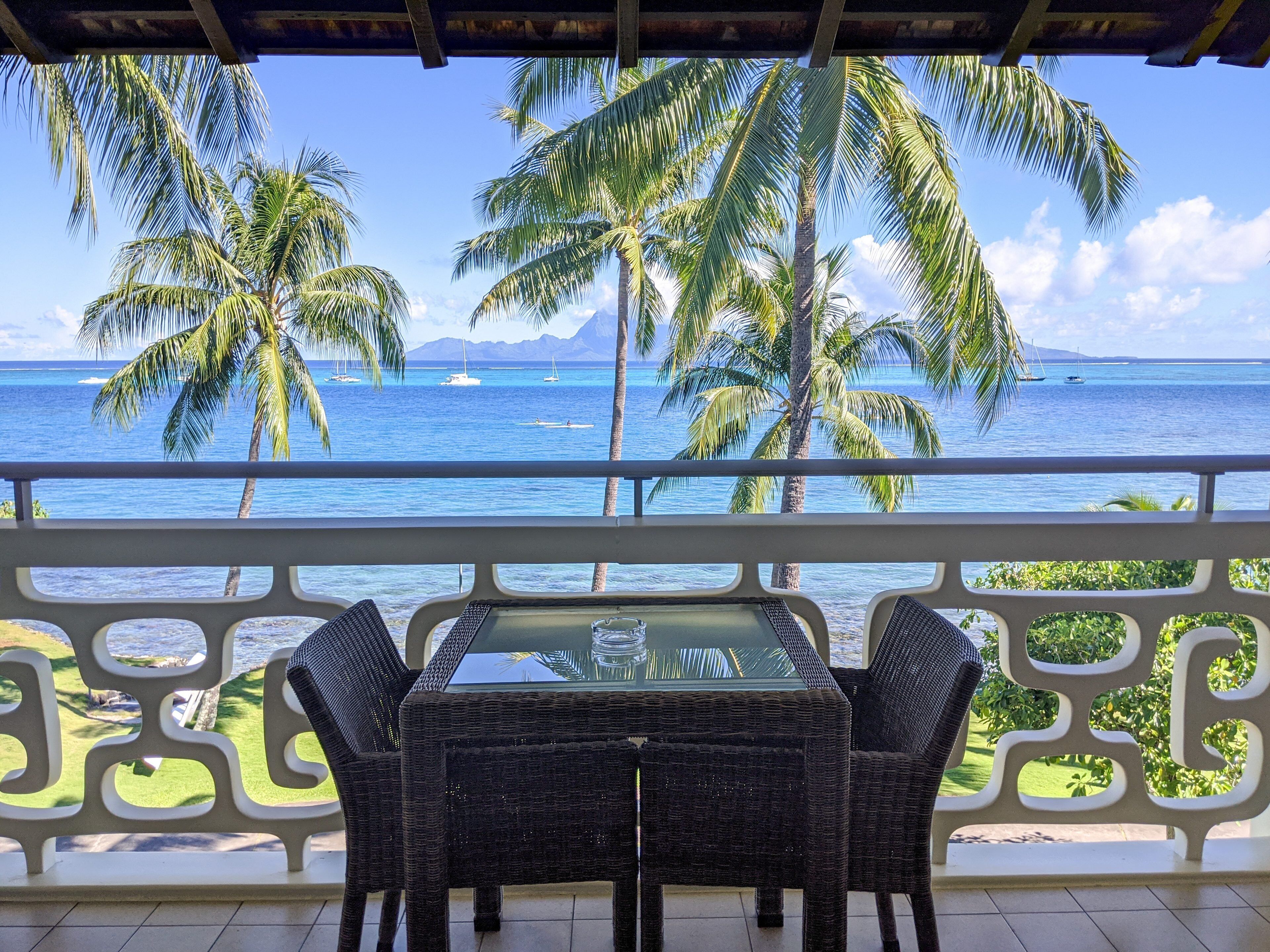 A balcony with wicker furniture overlooking a tropical beach with palm trees and boats.