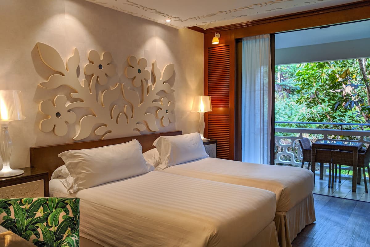A hotel room with two beds, a decorative wall, a balcony with a table and chairs, and a view of greenery.