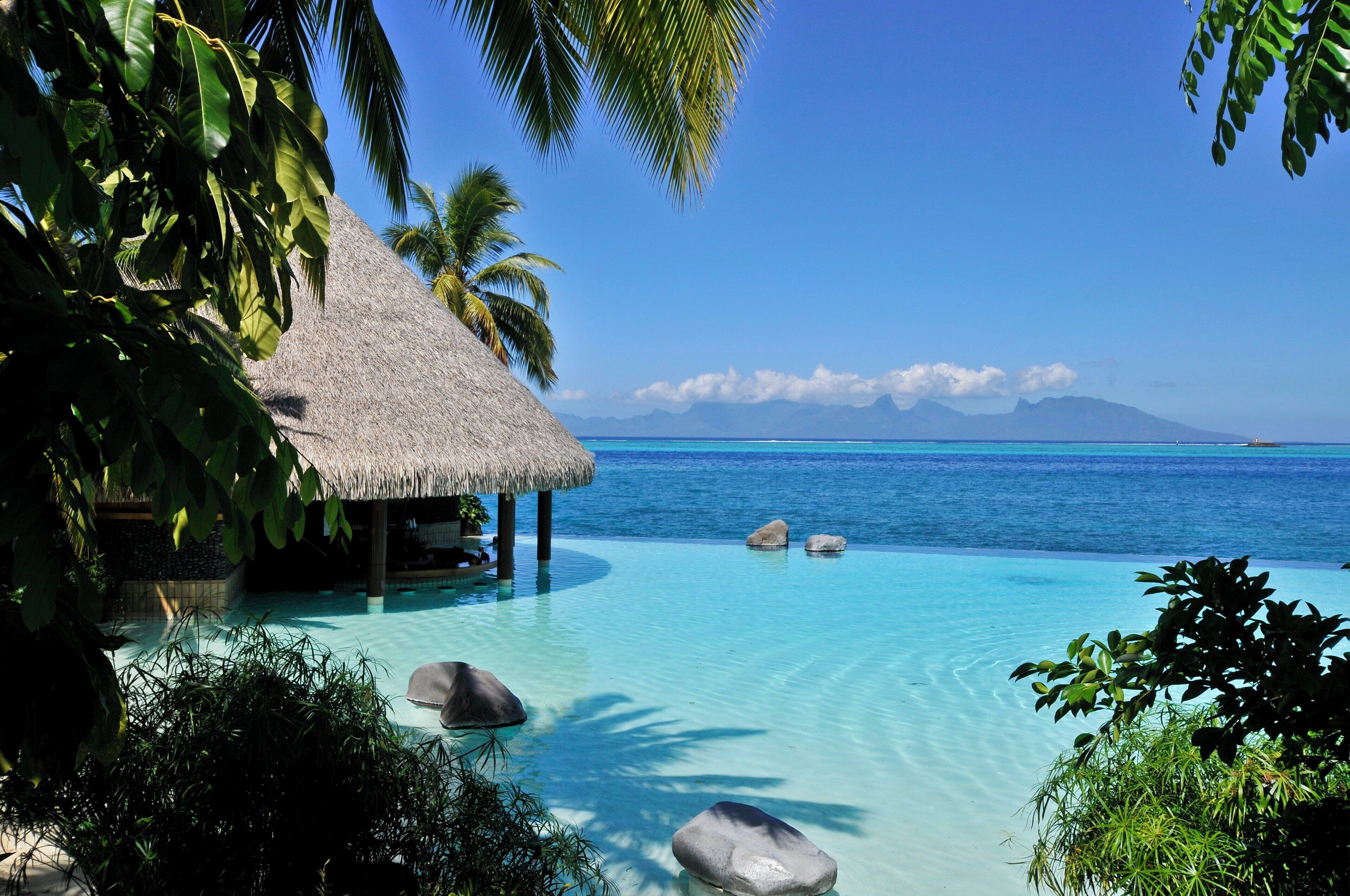 A clear tropical beach with a thatched-roof structure, calm turquoise waters, and distant mountains.
