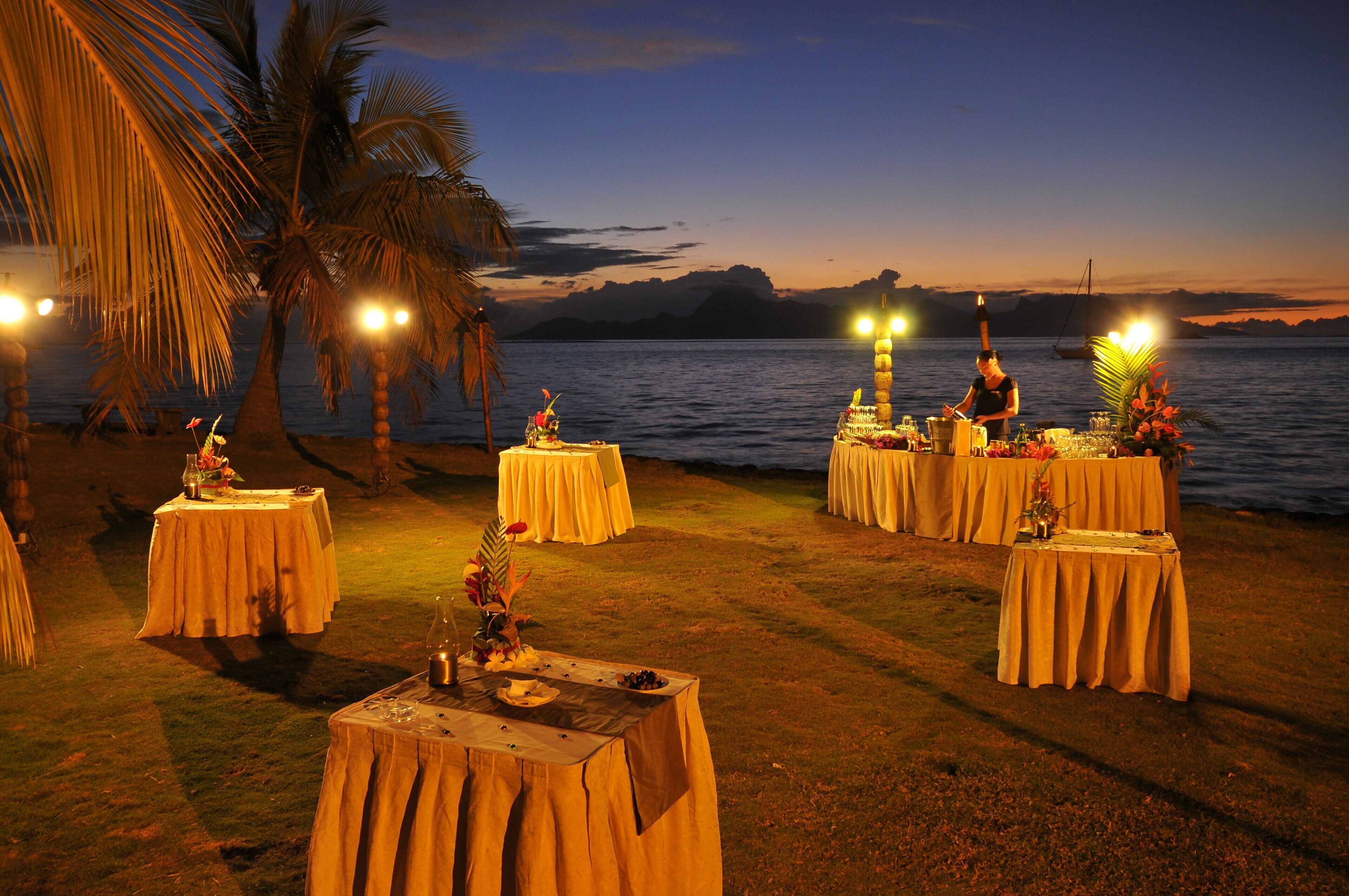 An outdoor dining area with tables set up on the grass near the water, palm trees, and a view of the water and mountains in the background.