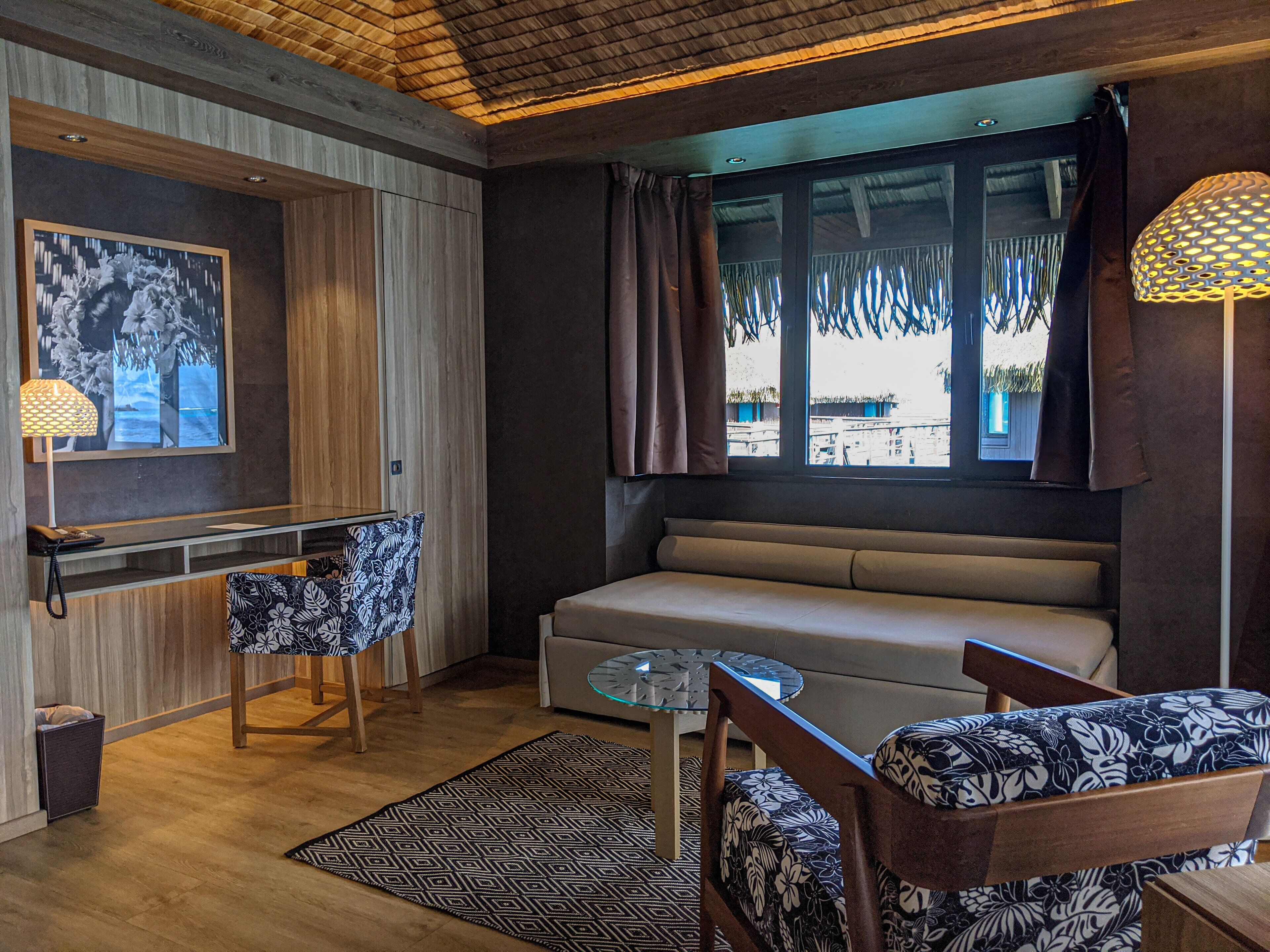 Una habitación moderna con sofá, una mesa de vidrio, sillas con estampado, un escritorio con lámpara y un ventanal con cortinas.