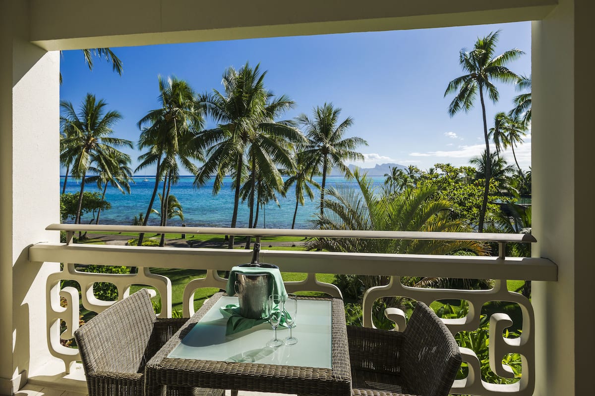 A balcony with wicker furniture, a table, and a view of the ocean and palm trees.