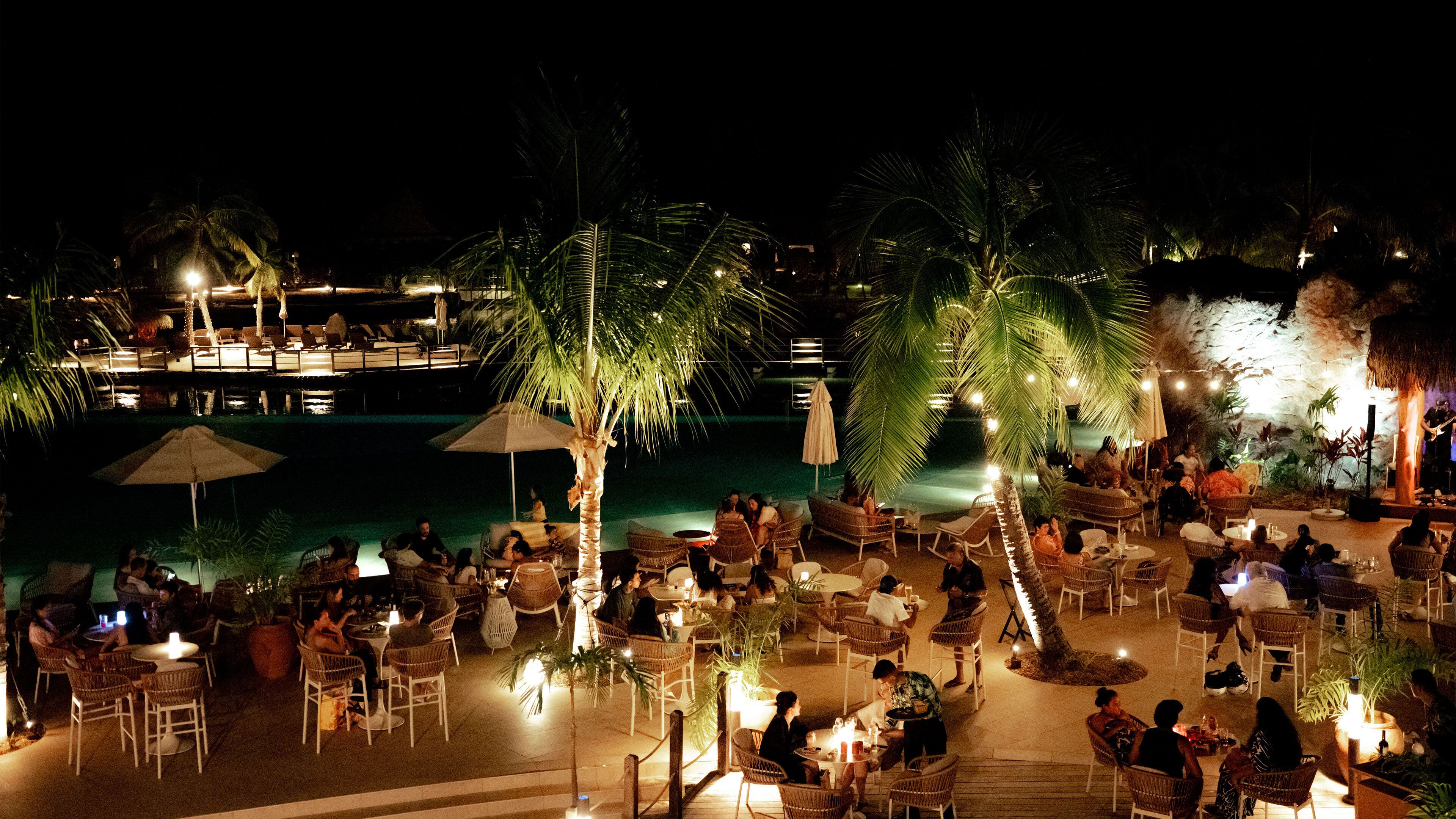 An outdoor dining area with tables and chairs, surrounded by palm trees and a pool.