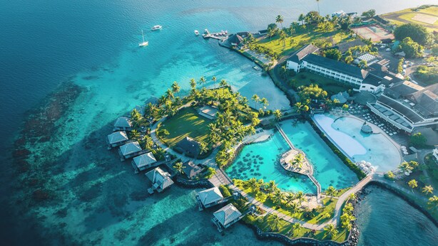 An aerial view of a resort with multiple overwater bungalows, a central pool area, and a large building likely serving as the main hotel.