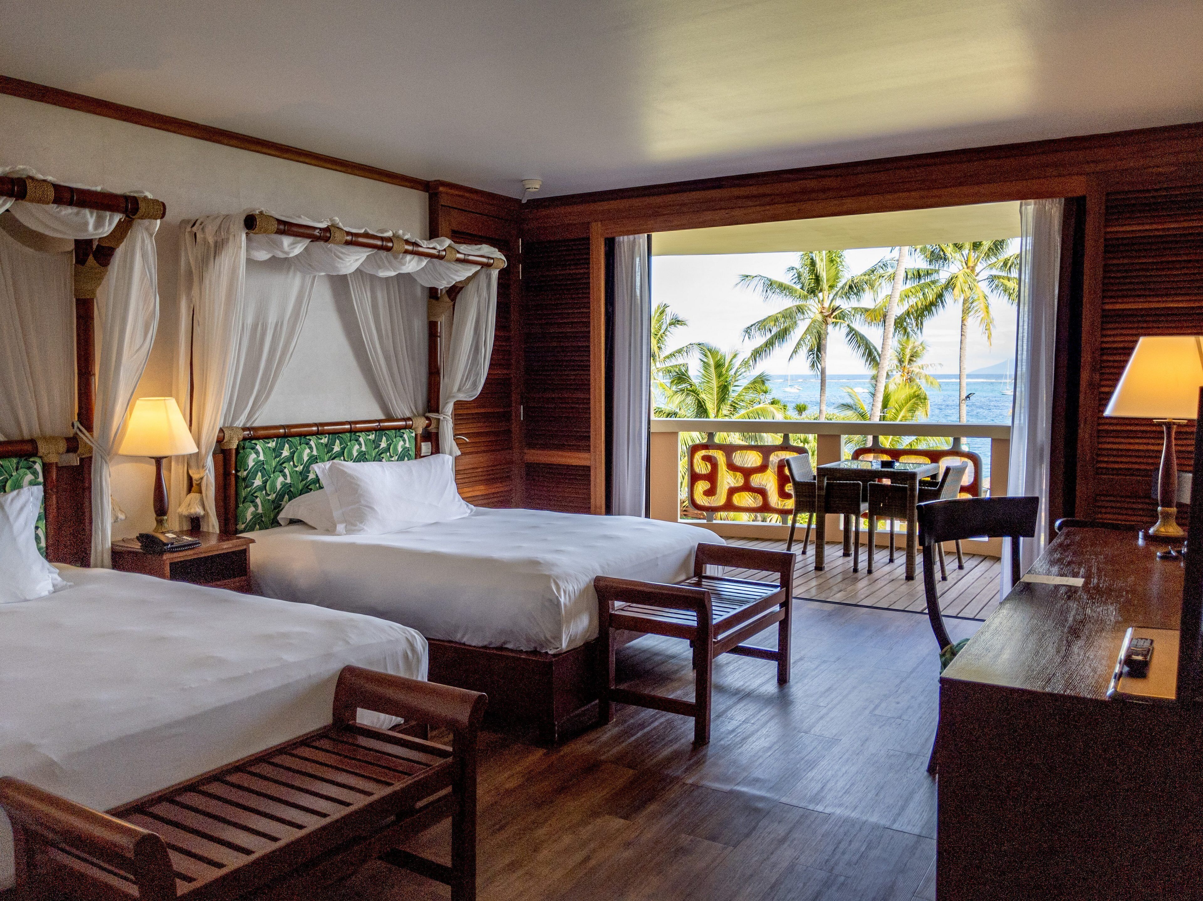 Une chambre d’hôtel avec deux lits, un balcon donnant sur des palmiers et l’océan, et un petit coin repas.