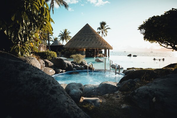 A pool area with a thatched-roof structure, surrounded by rocks and palm trees, overlooking a beach.