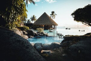 A pool area with a thatched-roof structure, surrounded by rocks and palm trees, overlooking a beach.