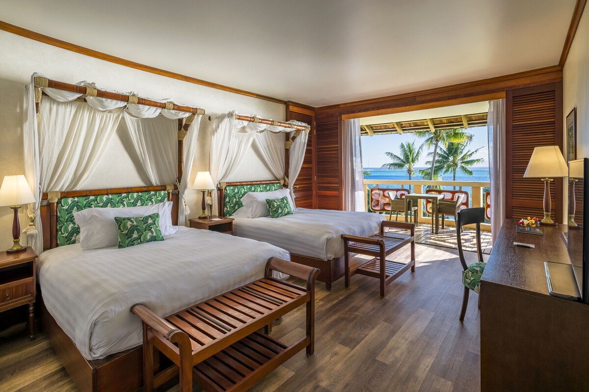 Une chambre d’hôtel avec deux lits, un banc en bois, un bureau et un balcon donnant sur l’océan.