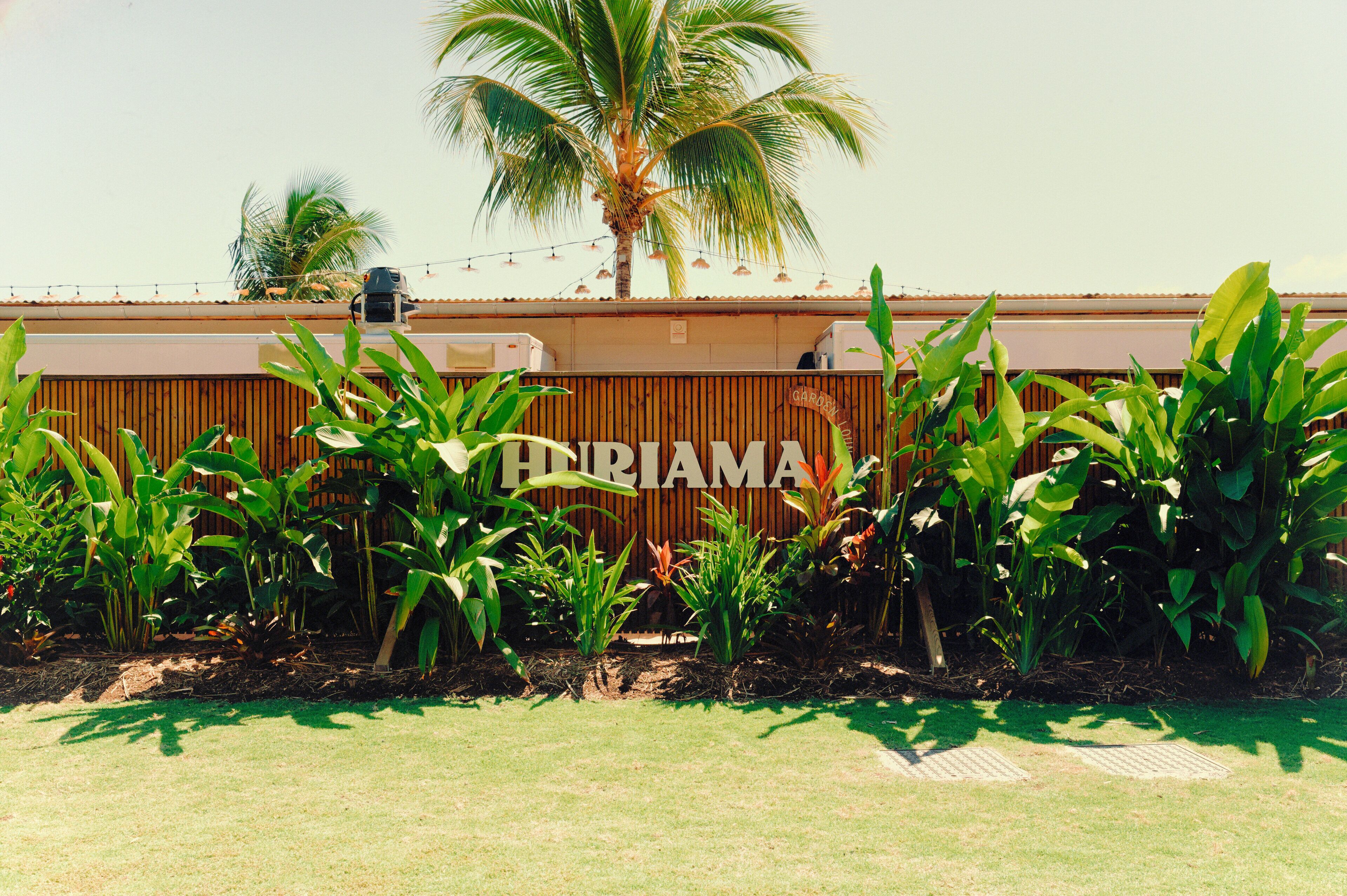 A sign with the name "HERRIAMA" on a wooden fence, surrounded by lush green plants and a palm tree in the background.