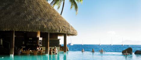A pool area with a thatched-roof structure, palm trees, and a view of the sea with sailboats.