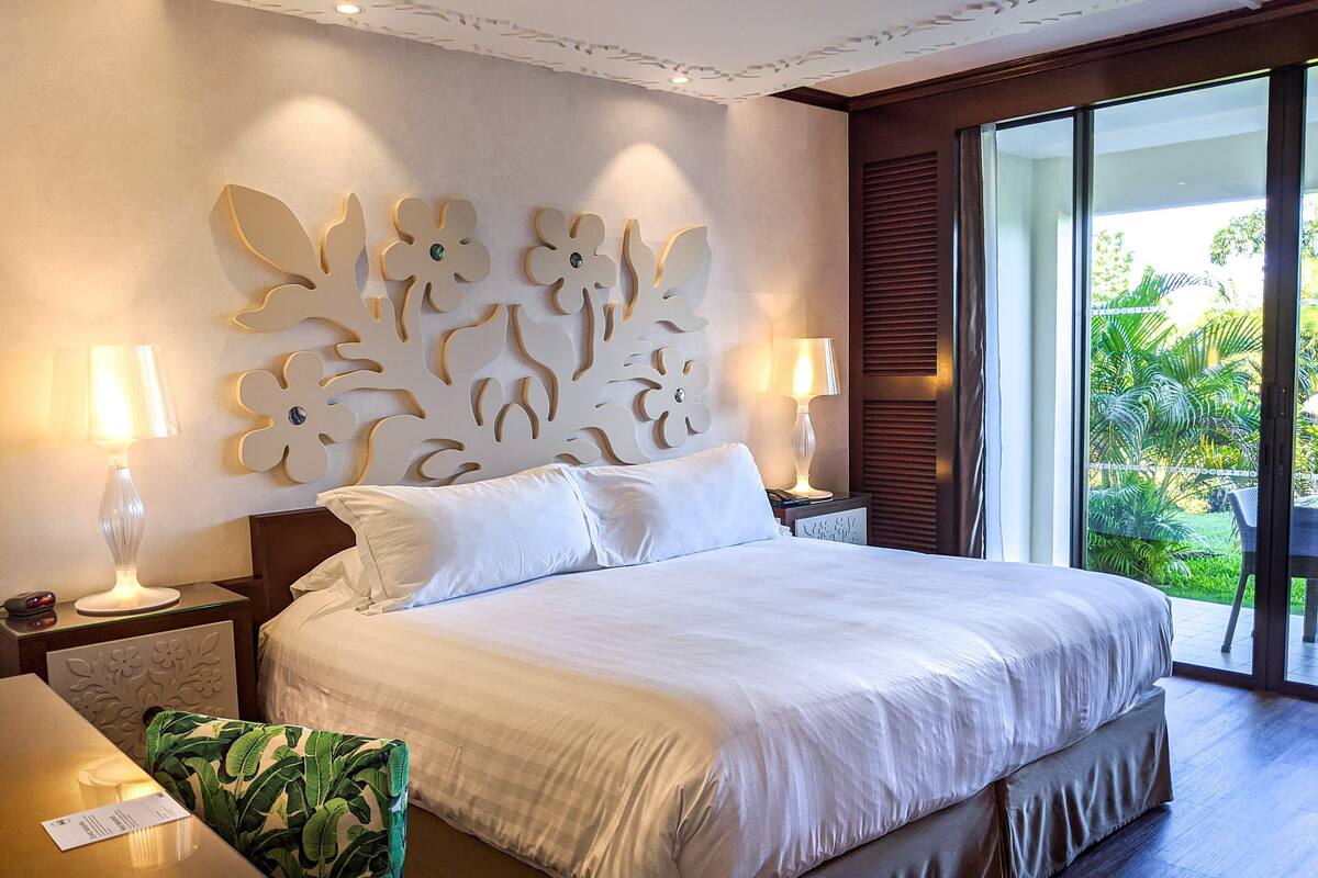 A hotel room with a large bed, two bedside lamps, a decorative wall panel, and a view of greenery through the window.