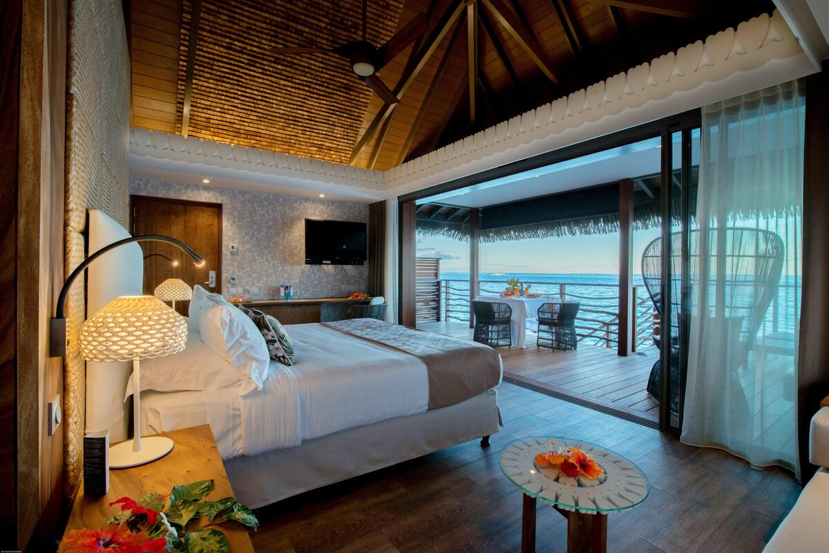 A bedroom with a bed, bedside table, lamp, and a view of the ocean.
