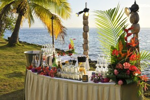 A table set up with drinks and flowers by the sea, with a decorative light fixture and a palm tree in the background.