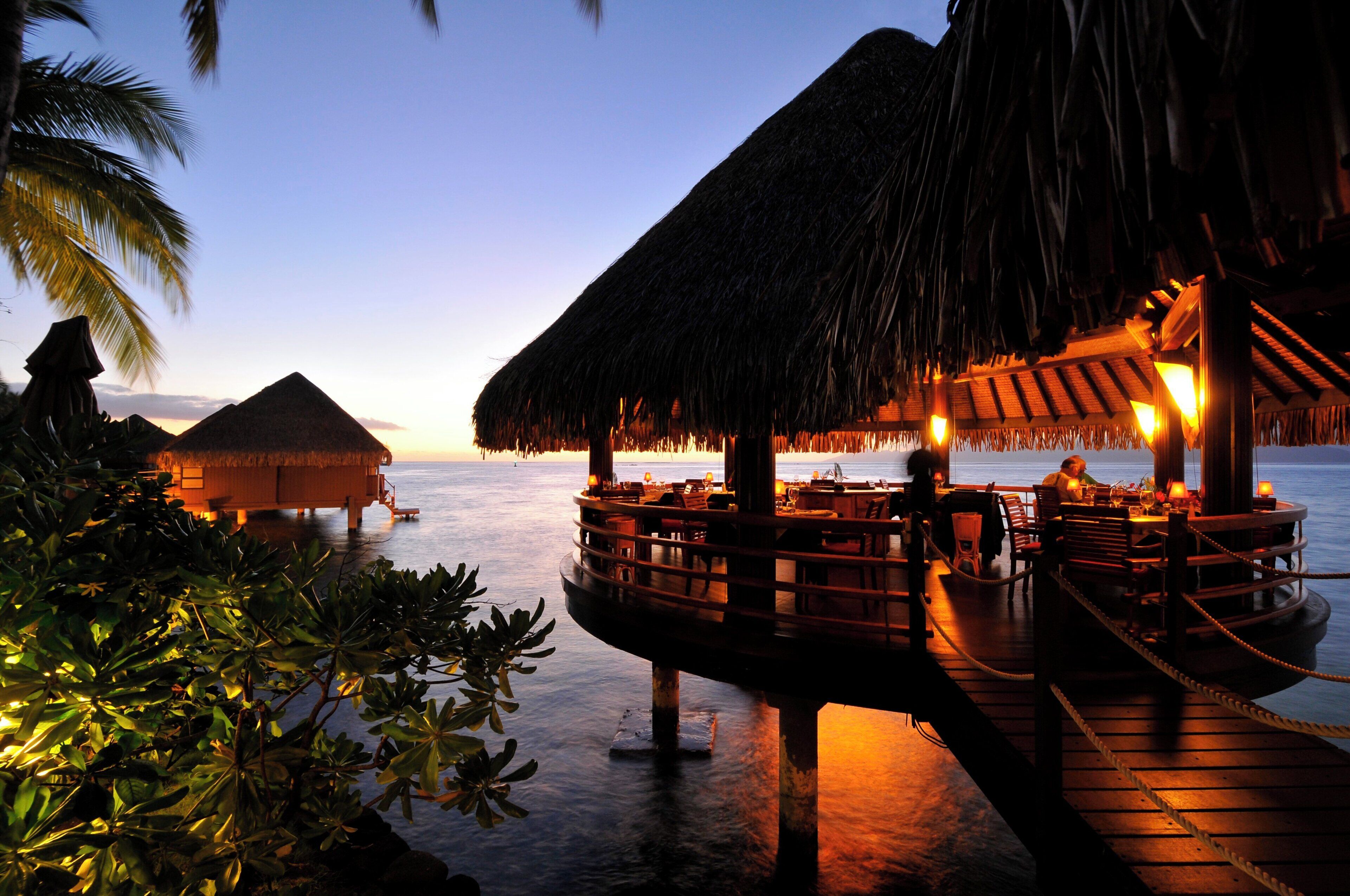 An overwater bungalows with thatched roofs, wooden deck, and dining area with tables and chairs.