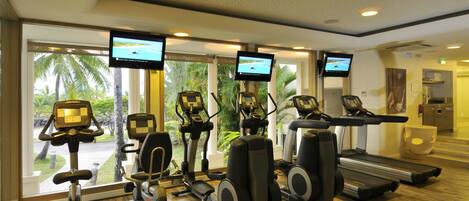 A gym with treadmills, elliptical machines, and stationary bikes, equipped with large windows and multiple screens.