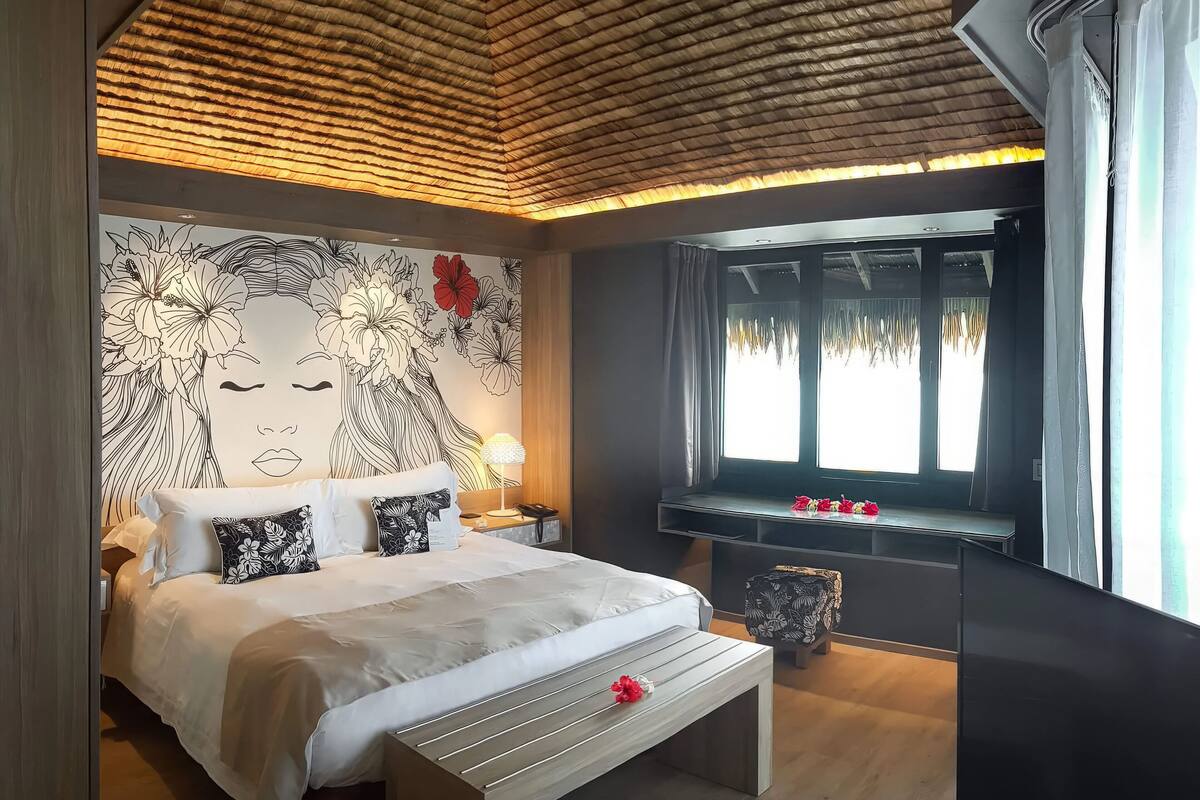 A modern bedroom with a large bed, a bedside table, a nightstand, a wall mural, and a window with curtains.