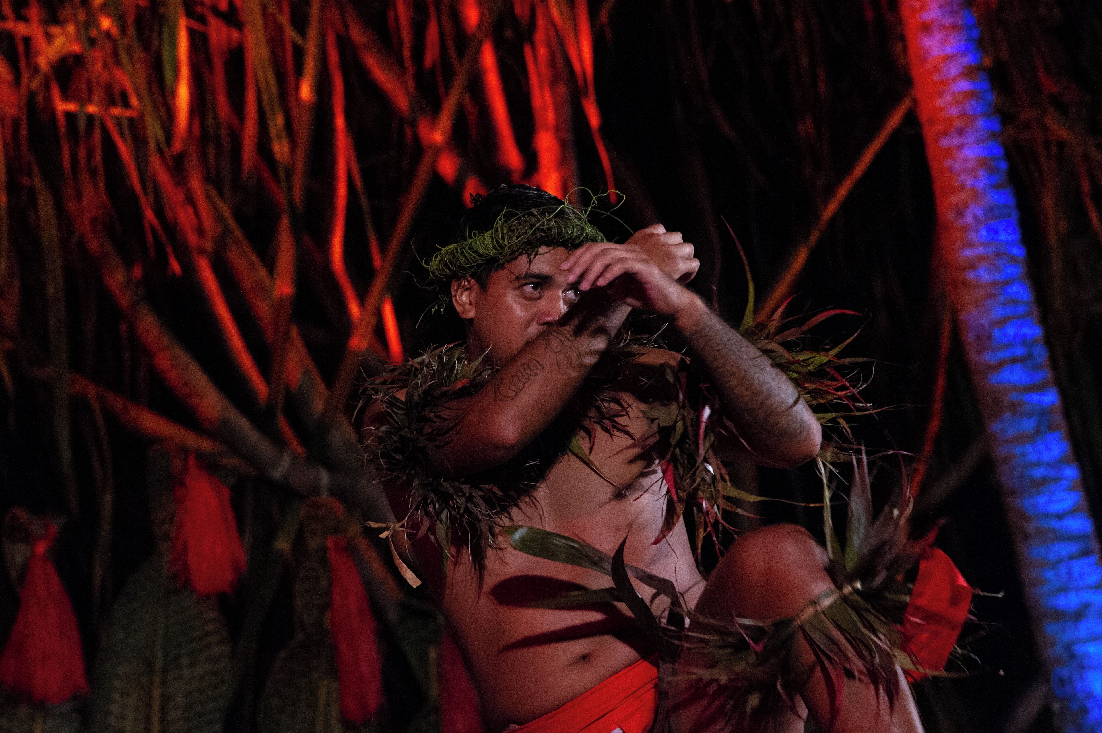 Uma pessoa vestida com traje tradicional, incluindo um headdress e uma vestimenta vermelha, cercada por elementos naturais como galhos e folhas.