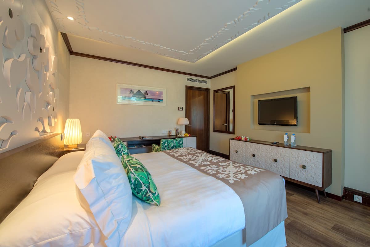 A hotel room with a large bed, a desk, a dresser, a TV, and a wall-mounted artwork.