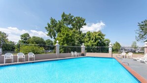 Ein rechteckiger Swimmingpool mit klarem, blauem Wasser, umgeben von einer niedrigen Mauer und einem Metallzaun. Weiße Plastikstühle stehen entlang des Poolrandes.
