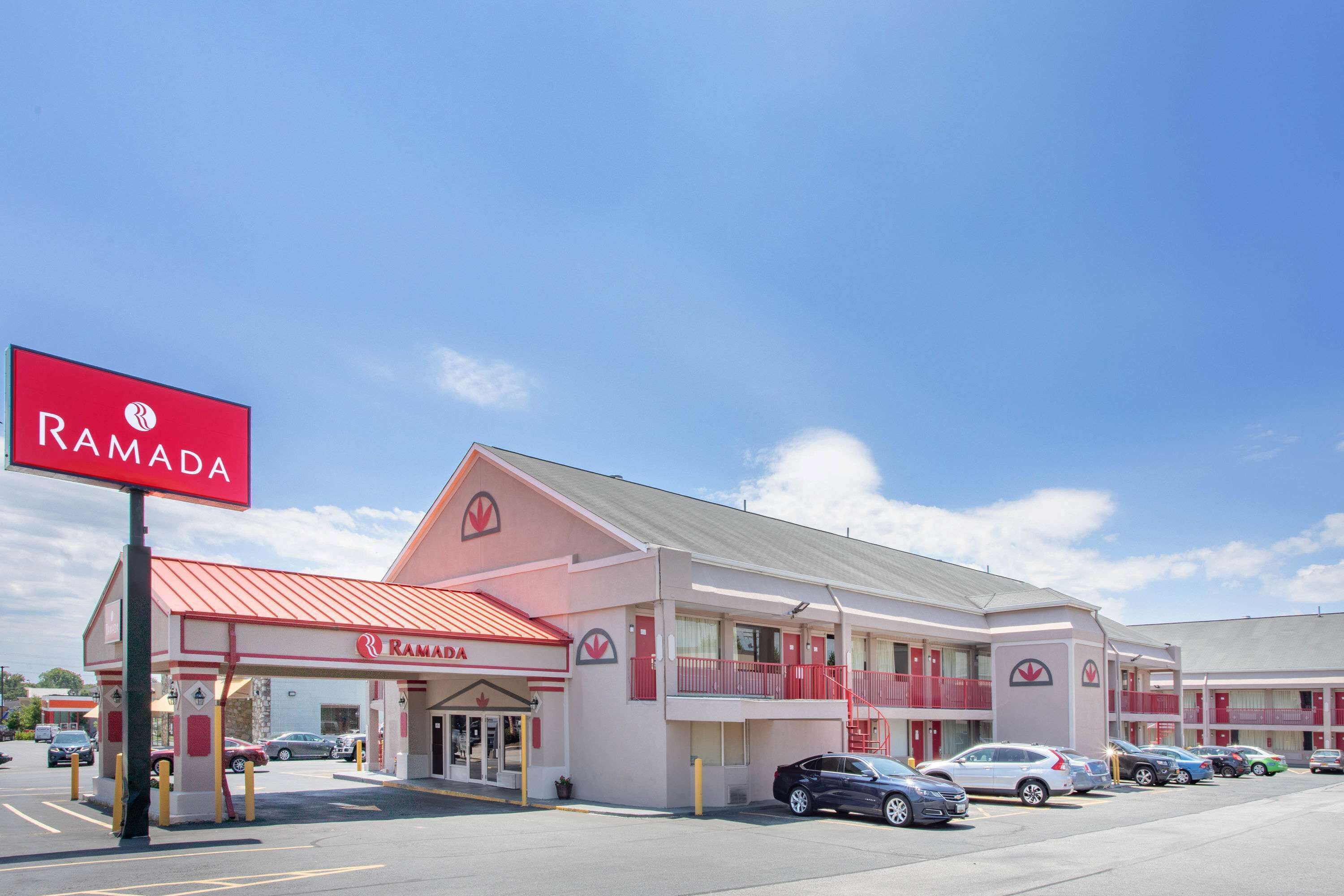 A Ramada hotel with a red and white facade, multiple balconies, and a parking lot with several cars.