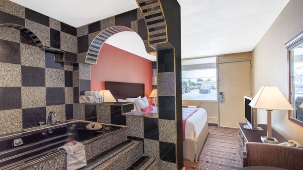 Ein Hotelzimmer mit karierter Fliesenwand, einem großen Spiegel, einem Bett mit rotem Kopfteil, einem Schreibtisch mit Lampe und Blick ins Freie durch eine Tür.