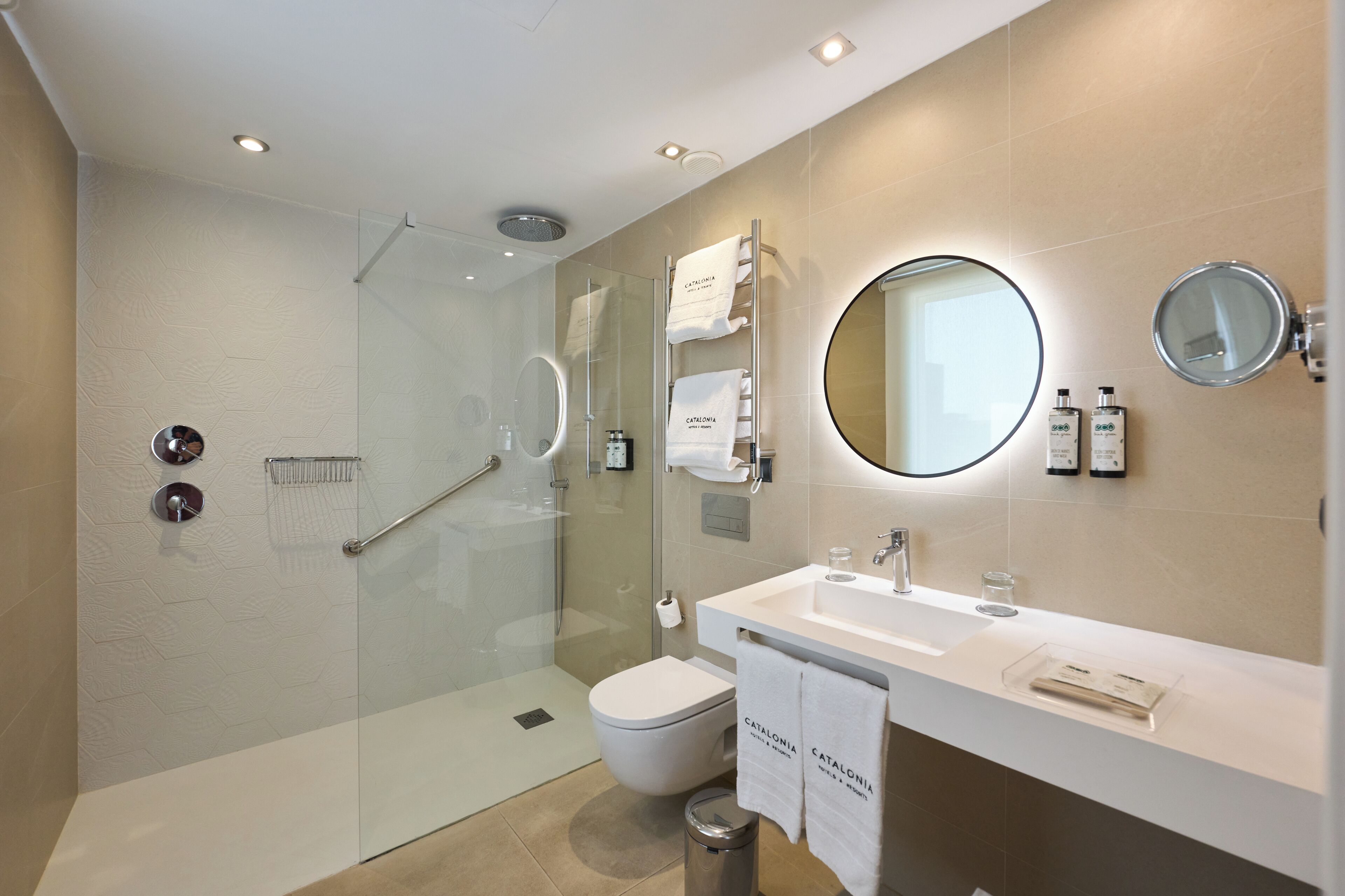 A modern bathroom with a glass-enclosed shower, a white sink, a toilet, and a round mirror.