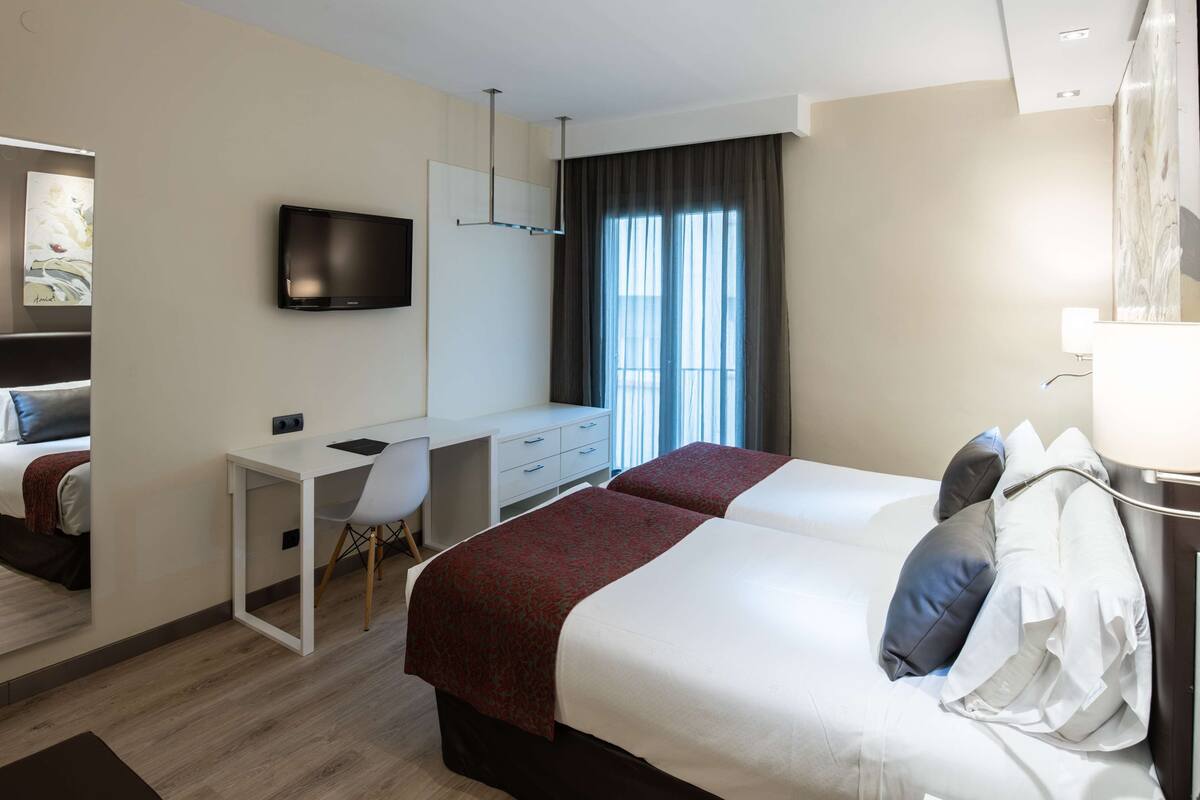 A hotel room with two beds, a desk, a chair, a television, and a window with curtains.