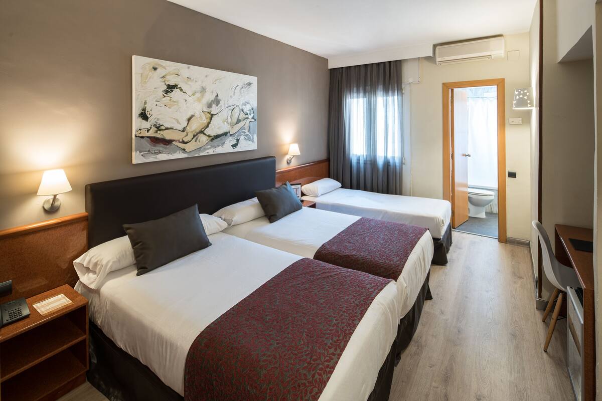 A hotel room with two beds, a desk, a chair, a lamp, and a painting on the wall.